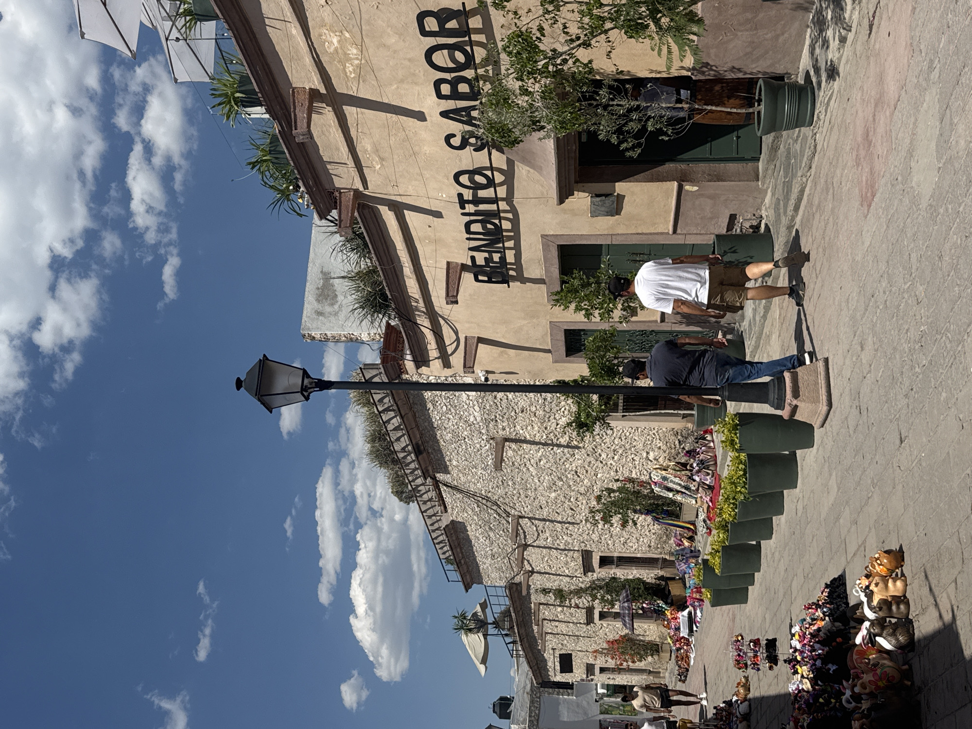 The scene depicts a street in Bernal, Mexico. Two pedestrians are visible: one wearing a white shirt and khaki shorts, and another in a blue shirt and jeans, walking on a stone-paved street next to a building. The building's facade displays the text "BENDITO SABOR" vertically. The building is constructed of stone and is adorned with plants in pots along its upper portion and near the entrances. A lamppost stands near the building. The street is lined with potted plants and merchandise for sale, including handcrafted animal figures. The sky above is partly cloudy.