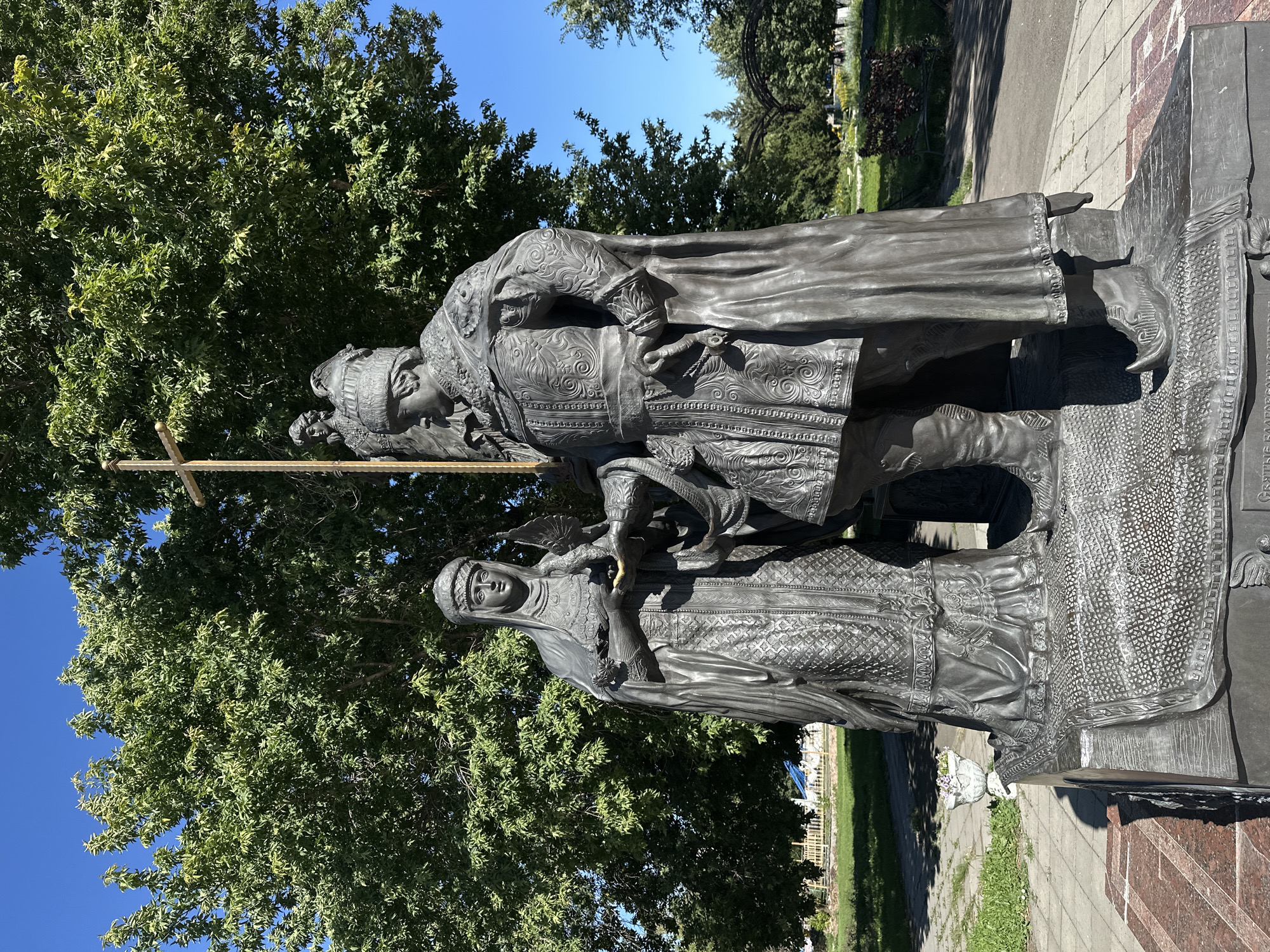 This image depicts a bronze sculpture of a man and a woman, likely historical figures, standing in a park on a sunny day. The man, on the right, is larger and wears ornate, medieval-style clothing and a fur-lined hat, holding a sword at his side. The woman, on the left, is dressed in a long, elaborate gown and head covering, holding a small bird in her hands. They are positioned on a sculpted base that appears to represent a rug or patterned surface. Behind them, a tall, golden cross or emblem rises against a backdrop of lush green trees and a clear blue sky. In the background, there are hints of park amenities like a bench and possibly some decorative arches. The sculpture is located in Biysk, Russia. The overall impression is one of historical commemoration within a pleasant, outdoor public space.