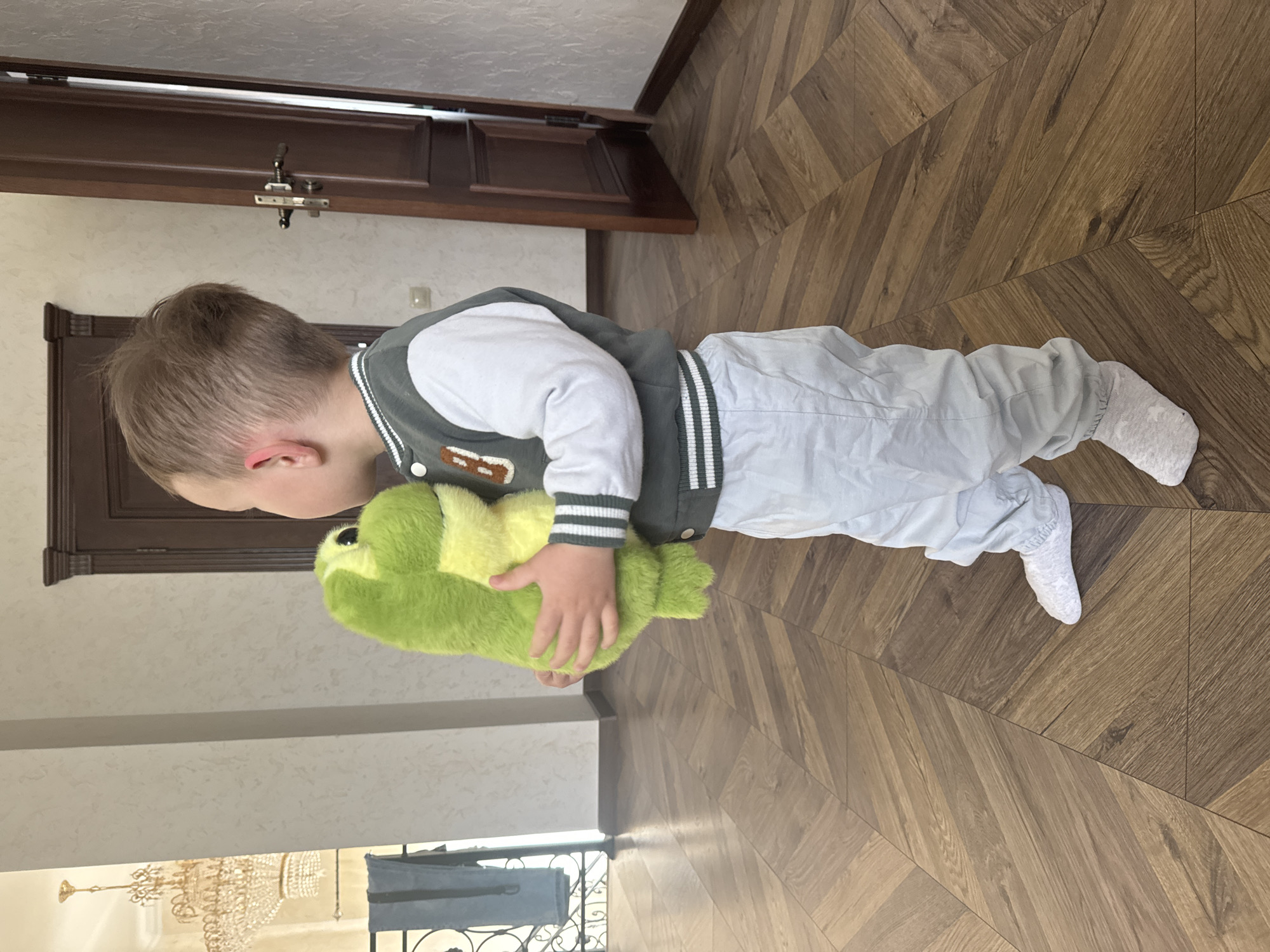 A child wearing a white long-sleeve shirt, gray pants, a gray jacket with white striped cuffs and collar, and white socks is standing on a wooden floor in a room. The child is holding a green stuffed animal that appears to be a frog. The background shows a doorway and a wall with a chandelier visible in the reflection. The image was taken in Макарово, Russia.