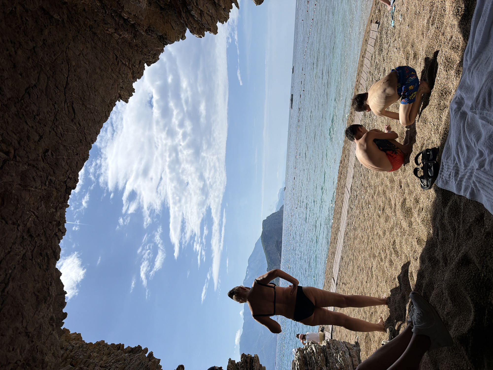 The image shows a beach scene in Budva, Montenegro. The perspective is from inside a rocky cave or overhang, framing the beach and sea. Two young boys in swim trunks sit on the beach to the right, interacting with something on the ground. A woman in a black bikini stands on the rocks near the cave entrance. Behind her, across the water, are mountains and a blue sky with streaks of clouds. The beach is sandy. The sea is calm.