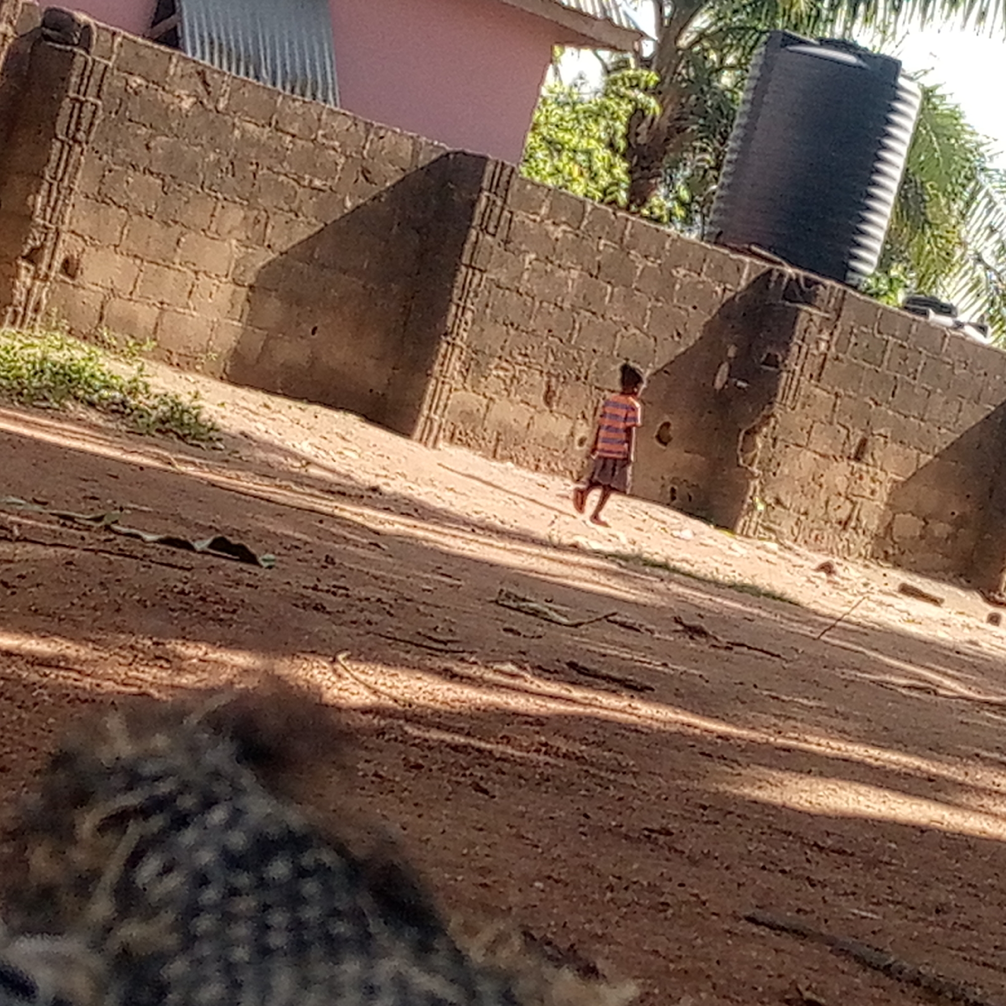 The image depicts a sunlit outdoor scene in Mararaba, Nigeria, focusing on a young child walking on a dirt path. The child, viewed from behind, wears a striped orange and blue short-sleeved shirt with dark shorts and is barefoot, moving towards the right side of the frame.
The ground is composed of reddish-brown dirt, with scattered lighter patches and some sparse green weeds along the edges. Long, clear shadows stretch across the path and up the wall, suggesting bright sunshine and either late morning or late afternoon.
In the background, a sturdy wall built from rough-hewn concrete blocks forms a prominent barrier. Behind this wall, a portion of a building with a light pink facade and a corrugated metal roof is visible. Green foliage, including a palm tree, can be seen peeking over and behind the wall. A large, black cylindrical water tank stands on top of the wall or a structure behind it, on the right side of the image.
The bottom-left foreground contains an out-of-focus, dark, and textured element. No discernible text is visible in the image. The scene conveys a calm, everyday moment in a warm, bright environment.