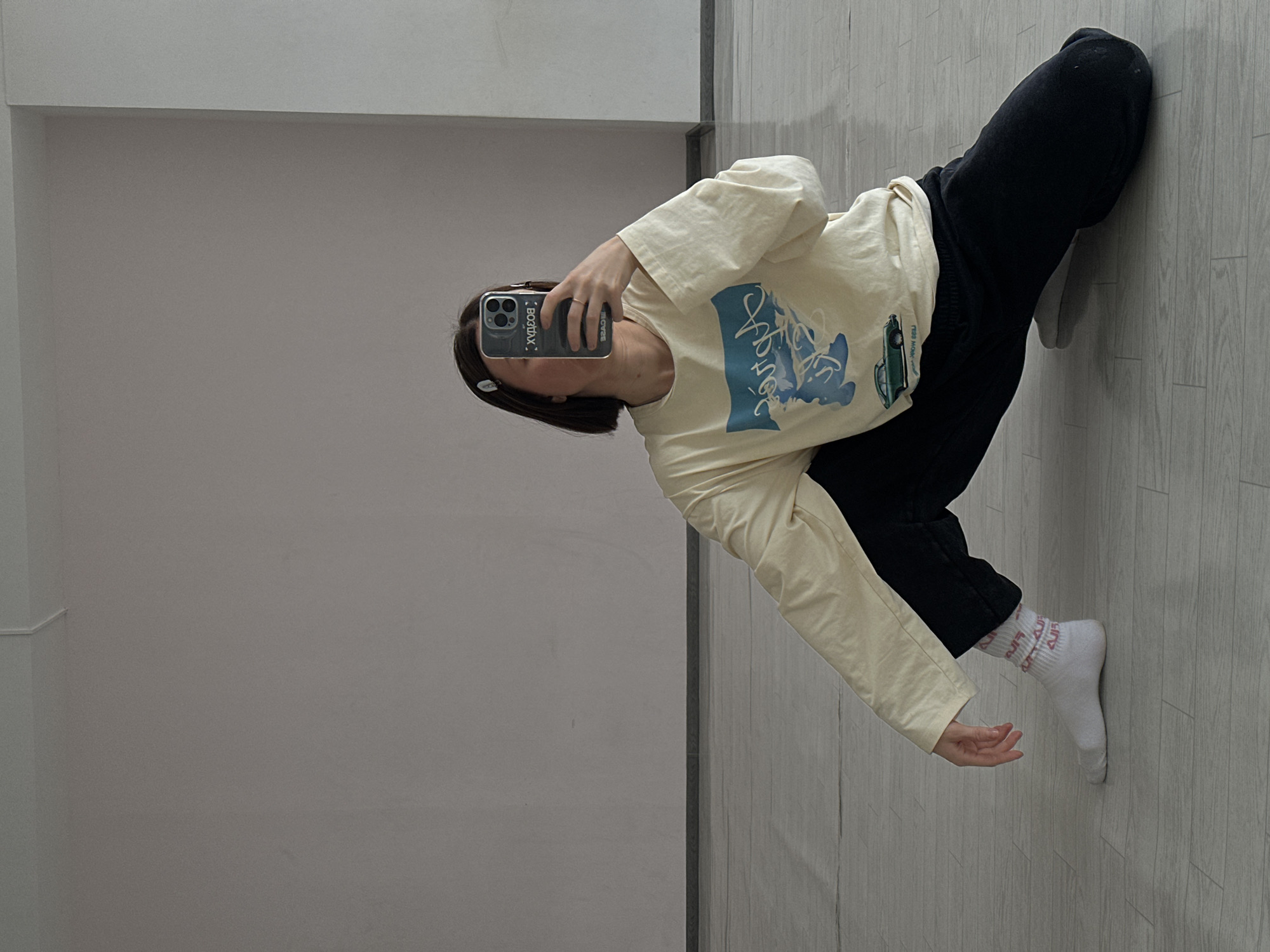 A woman is taking a selfie indoors. She's wearing black pants, a light yellow long-sleeved shirt with a blue design, and white socks with pink lettering. The woman is holding a smartphone in her left hand to take the photo. The background consists of a light gray wall on the left and a light gray wooden floor on the right. There is no indication of location in the city of Votkinsk, Russia.