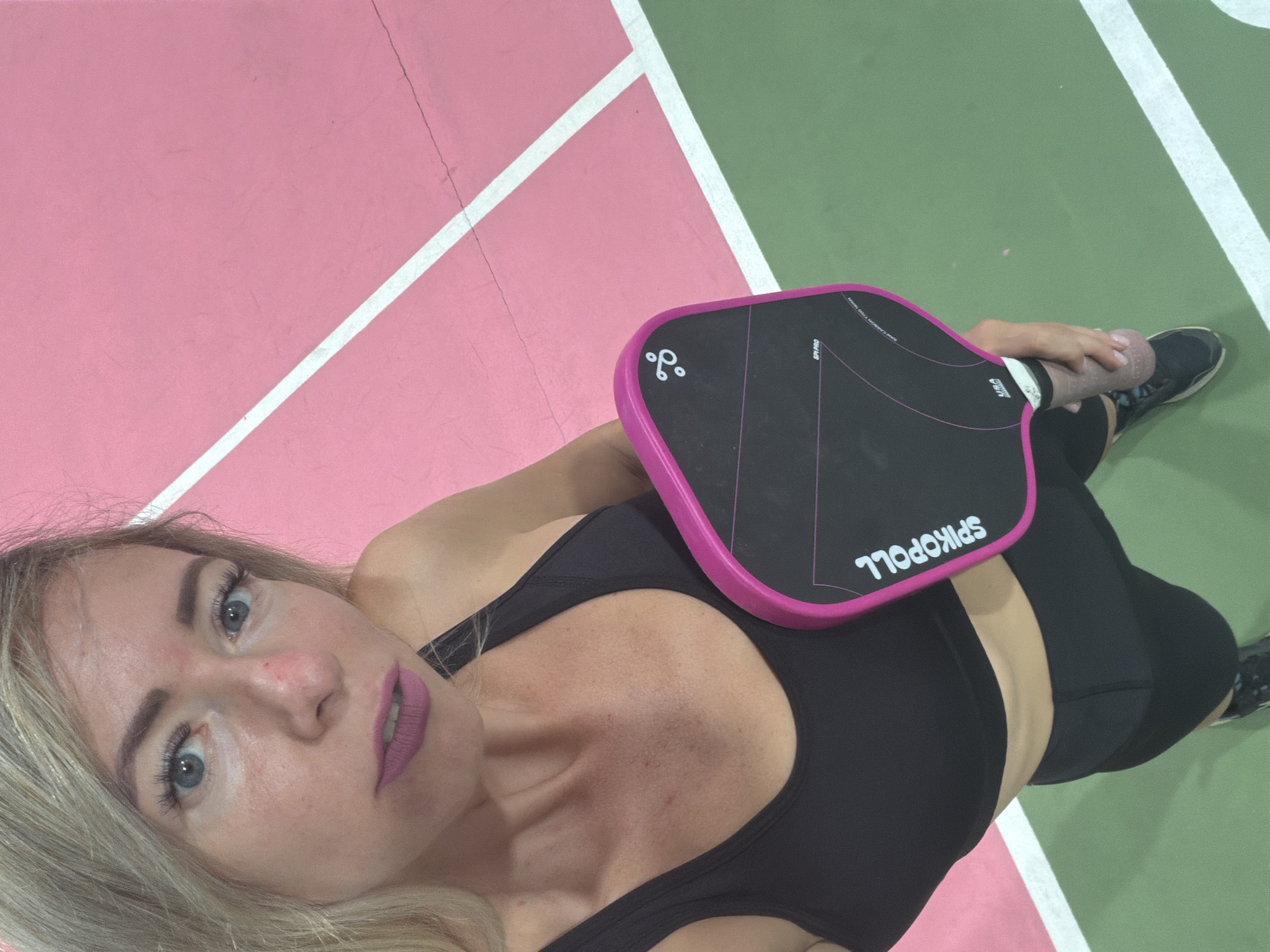 A young woman with blonde hair and blue eyes, wearing a black sports bra and black shorts, holds a pink and black pickleball paddle. She is standing on a pickleball court with pink and green surfaces and white lines. The image is taken from a high angle, looking down at the woman. She appears to be posing for a selfie. The location is not specified in Da Lat, Vietnam.