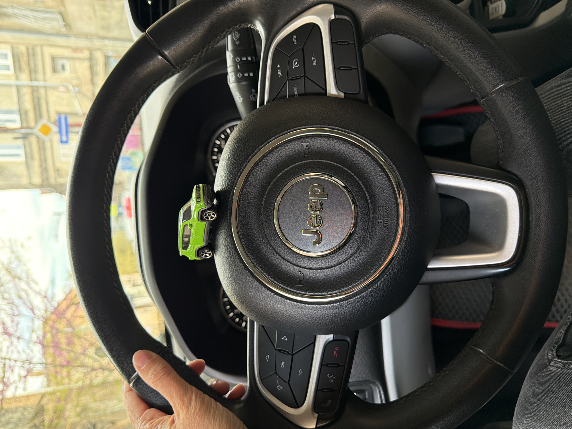 The image presents an interior perspective from inside a car, focusing on the steering wheel and the view through the windshield. A human hand, likely an adult's, firmly grips the lower left portion of a black leather steering wheel. The steering wheel is adorned with silver accents and integrated controls, including buttons labeled "RES," "SET+", "SET-," "CANC," "OK," along with various arrow and phone icons for media and cruise control functions. The prominent "Jeep" logo is centered on the horn/airbag cover, with "SRS AIRBAG" partially visible beneath it. A small, vibrant green toy car, resembling a Jeep Wrangler, is attached as a decorative element to the left spoke of the steering wheel.
Behind the steering wheel, a part of the dashboard and instrument cluster is visible, along with a wiper/turn signal stalk on the left. On the lower right, a person's leg, dressed in dark denim jeans, and a grey shoe are present.
Through the windshield, a daytime urban environment in Tbilisi, Georgia, is visible, albeit slightly blurred. The background features multi-story buildings, street signs (including a distinctive blue and yellow diamond sign), and trees with sparse branches, indicating late winter or early spring. The natural lighting suggests clear weather conditions. The scene depicts a driver holding the steering wheel, possibly paused or in transit within the city.