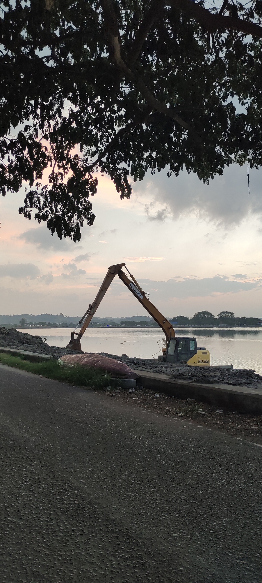The image depicts a waterfront scene in Lhokseumawe, Indonesia, likely during twilight hours. The foreground features a paved road with a grassy verge bordering a body of water. On the embankment beside the water, a yellow excavator is positioned, its boom extended upwards and outwards over the water. The excavator appears to be engaged in some form of construction or dredging activity, with piles of dirt or debris visible on the bank. The water reflects the cloudy sky, which has hints of pink and orange, suggesting a sunset or sunrise. In the background, a distant shoreline is visible with trees and buildings, indicating a settled area. Overhanging the scene from the top of the image is the dark silhouette of a large tree with dense foliage, casting shadows and framing the sky and water. No people are visible in the image.