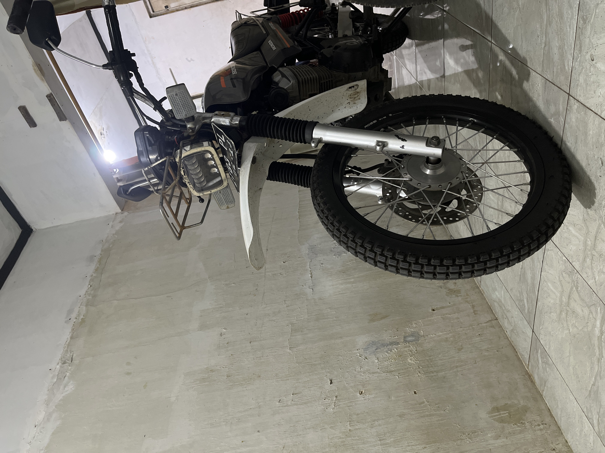 The image shows a black and white Honda motorcycle parked indoors, likely in a garage or workshop in Pekalongan, Indonesia. The motorcycle is positioned facing forward and slightly to the right, with its front wheel prominently in the foreground. The wall behind the motorcycle appears to be roughly plastered and painted a light color, showing some signs of wear and tear. The floor is tiled with a light-colored, marble-patterned tile.
The motorcycle itself is a dual-sport or scrambler type, evident from its knobby tires, high-mounted front fender, and exposed engine components. It has a rectangular headlight with multiple LED elements, a small front rack above the headlight, and turn signals mounted on either side. The fuel tank is black with orange and white "Honda" branding. The rear suspension is visible with a red spring. The handlebars are black and equipped with mirrors. A license plate, partially obscured, is mounted below the headlight. The overall condition suggests it is either a well-used or modified motorcycle. The lighting in the scene appears to be artificial, casting shadows on the floor.