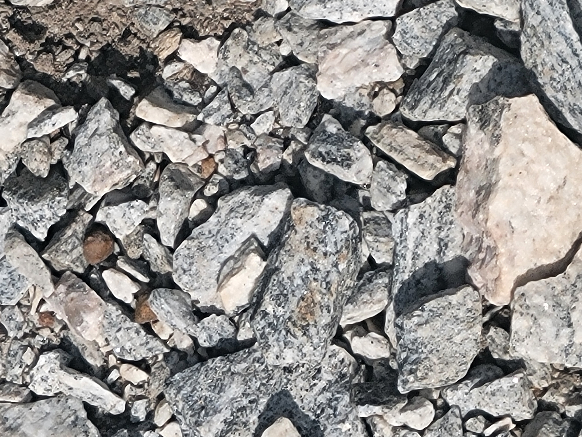 The image displays a close-up, overhead view of a pile of rough, angular gravel and stones. The stones vary in size, with some pieces being small and granular, while others are larger, irregular chunks. The dominant colors are shades of gray, white, and some hints of brown and tan. The textures appear rough and mineral-like, with visible crystalline structures on some of the larger stones. Shadows are cast across the gravel, suggesting an outdoor setting with directional lighting. The scene is devoid of any human presence or identifiable landmarks within the frame. Based on the visual information, it is not possible to determine the specific location within the city of Ogbomosho, Nigeria, as there are no discernible contextual elements.