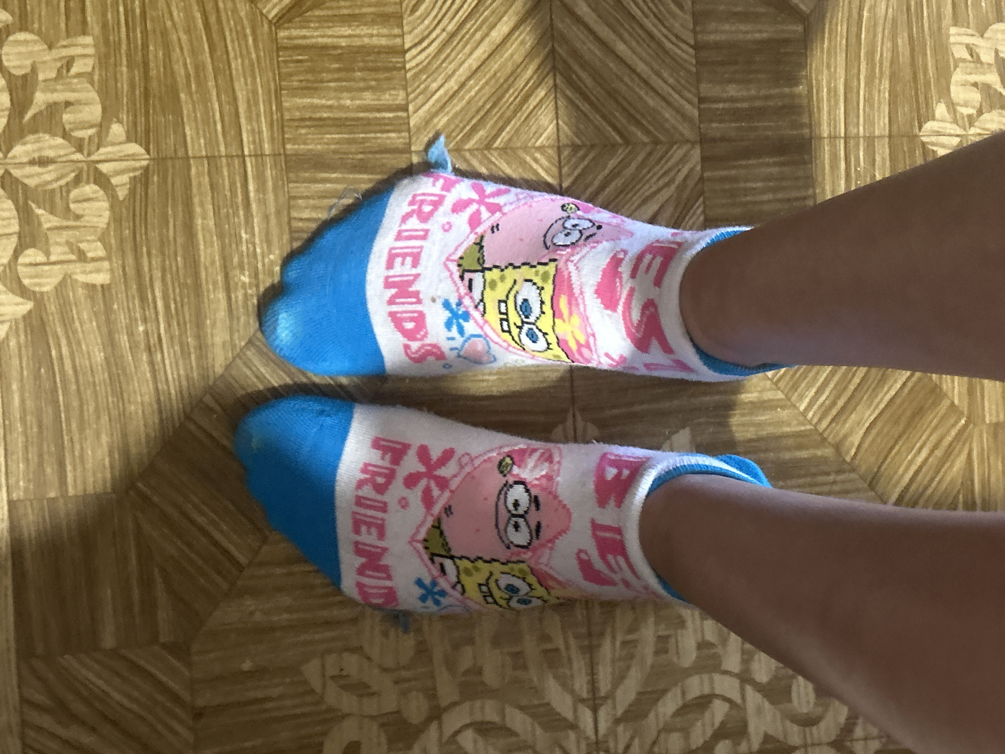 The image shows a pair of white ankle socks with blue cuffs, each featuring cartoon characters SpongeBob SquarePants and Patrick Star. The socks are designed to be worn by "best friends," as indicated by the text "FRIENDS" printed in pink on the cuff of one sock and "BEST" visible on the cuff of the other. The socks are worn by a person whose legs are visible, standing on a wooden-patterned floor. The floor appears to be linoleum or tile with a wood grain design, and it has a floral or decorative pattern in lighter beige tones. The lighting suggests an indoor environment. There is no visible information in the image that links it to Perushtitsa, Bulgaria.