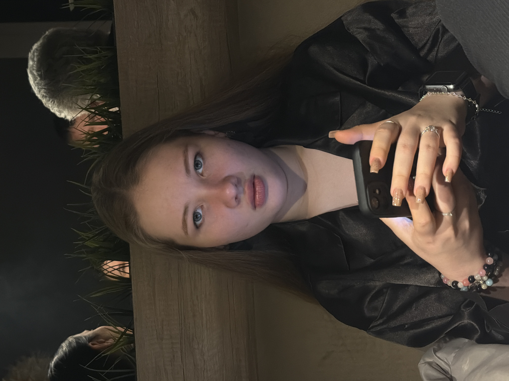 A young woman with long brown hair and blue eyes is holding a smartphone, possibly taking a selfie or using it. She is wearing a dark, possibly satin, shirt and several rings on her fingers, along with a watch and bracelets. She appears to be seated at a table or counter made of light wood. Behind her, partially obscured by artificial grass, are glimpses of other people. The location might be Chaikovsky, Russia, as specified in the prompt.