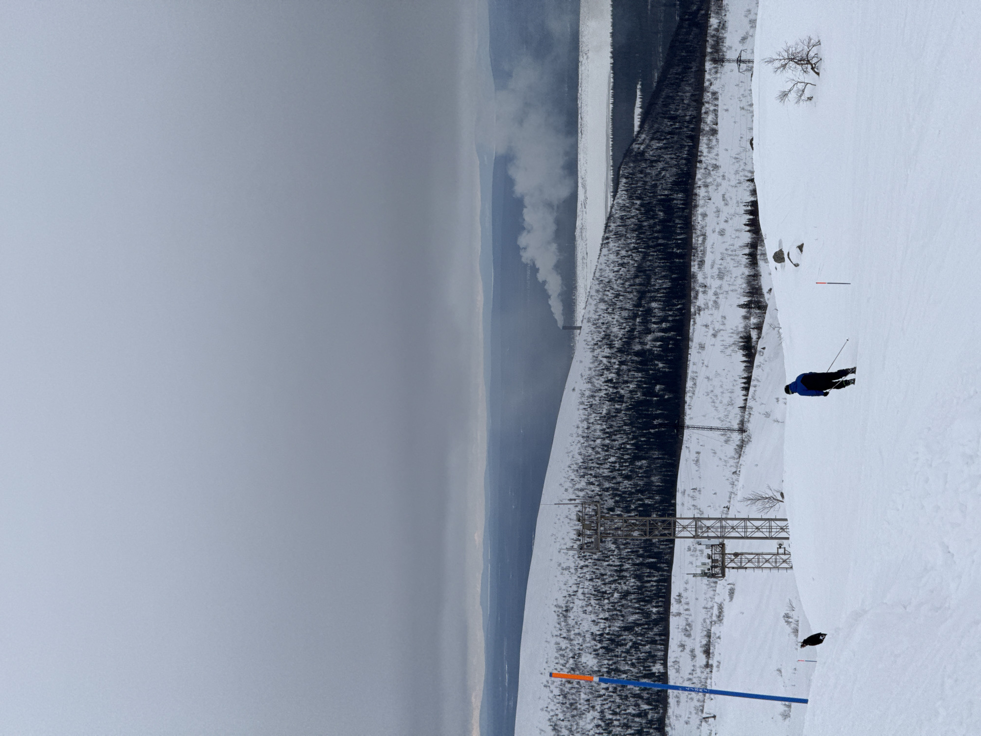 The image depicts a panoramic winter scene on a snow-covered mountain slope in Kirovsk, Russia, likely part of the Bolshoy Vudyavr ski area. The sky is overcast and grey. In the foreground, a lone skier, dressed in a dark blue jacket and dark pants, descends the pristine white slope towards the right, holding ski poles. A second individual in dark clothing is visible higher up the slope to the left, standing or walking. A blue marker pole with an orange top is positioned on the far left of the foreground, and a smaller orange marker pole is further right. Sparse, snow-dusted vegetation and a few rocks are scattered across the immediate slope.
In the midground, a prominent metal lattice tower, equipped with various antennas, stands on a snow-covered rise, surrounded by a dense, dark coniferous forest line. Further back, another smaller power line tower is visible. The background reveals a wide, partially frozen and snow-covered body of water, presumably a lake, bordered by dark forest. Beyond the lake, an industrial complex is identifiable by a tall, slender smoke stack emitting a substantial white plume of smoke that drifts horizontally across the landscape. Distant, snow-covered mountains and hills complete the horizon under the grey sky.