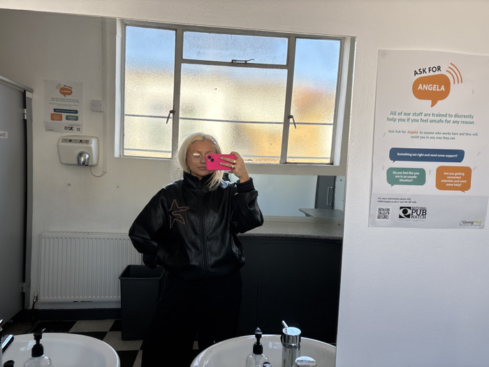The image displays a fair-skinned female capturing a mirror selfie inside a public restroom, likely located within a pub or bar in London, United Kingdom. This specific location inference is supported by the prominent "ASK FOR ANGELA" safety poster on the right wall, which includes "NATIONAL PUBWATCH" and "GivingPoint" logos. The individual, characterized by blonde hair and glasses, wears a black jacket with a light-colored star outline on the left chest and dark trousers. She holds a pink smartphone, partially obscuring her face, to take the photograph. In the foreground, two white ceramic sinks are visible, equipped with chrome faucets and soap dispensers. The left wall features a white hand dryer mounted above a white panel radiator, adjacent to a black waste bin. A large, multi-pane window in the background provides a blurred view of an outdoor scene under a blue sky. The room exhibits white painted walls, a black and white checkered tile floor, and a grey door partially visible on the far left. A second, partially obscured safety-related poster is present on the left wall.