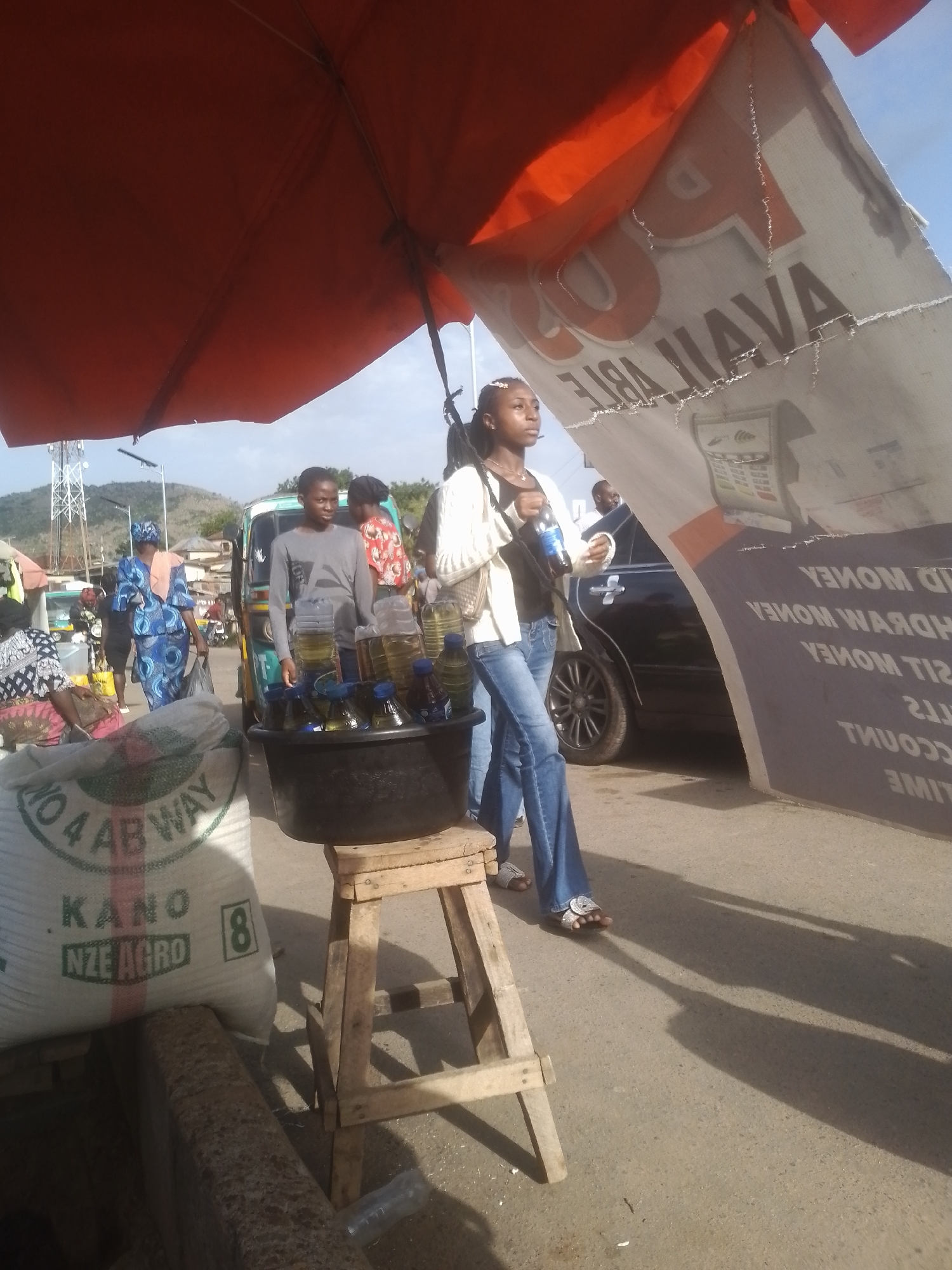 A bustling outdoor market scene unfolds under a large orange umbrella. A young woman in a white top and blue jeans stands near a stall, holding a bottle of water. In the foreground, a large sack with "KANO NZE AGRO" printed on it is visible. A black basin filled with bottles of yellow liquid, likely oil or fuel, sits on a wooden stool. Behind the stall, a man in a grey long-sleeved shirt stands next to a light-green auto-rickshaw. Further in the background, people are walking and interacting amidst market stalls. The environment appears to be a street with a paved surface, and in the distance, there are hills and a tall tower. The lighting suggests it is daytime.