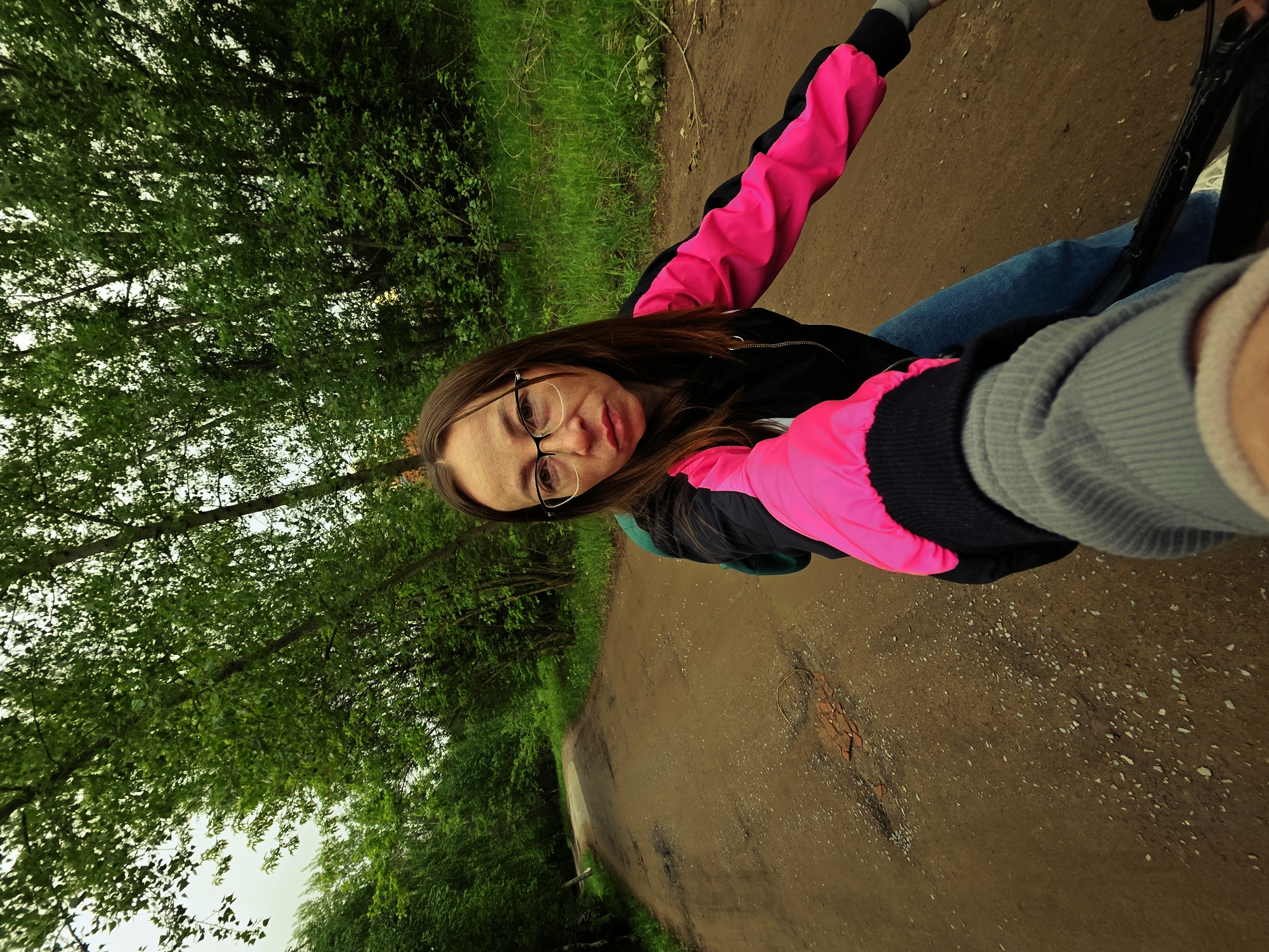 A woman with glasses is taking a selfie while riding a bicycle on a dirt path. She wears a black jacket with pink sleeves and blue jeans. The background shows lush green trees and vegetation. The location is in the city of Chaikovsky, Russia, as determined by the location metadata. The path is unpaved and appears to be a trail through a wooded area.