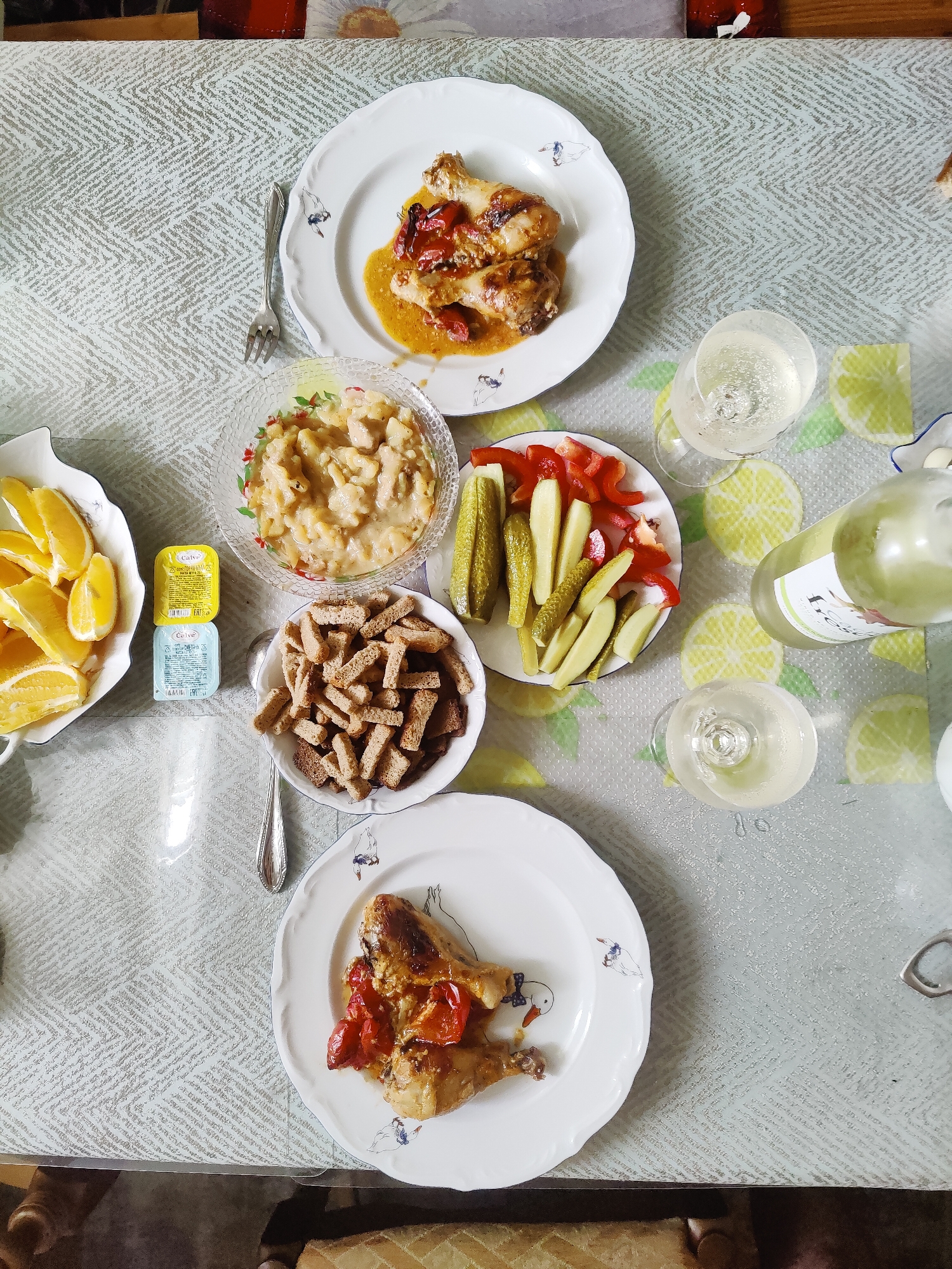 An overhead shot shows a meal laid out on a table. The table is covered with a tablecloth with a geometric pattern. On the table, there are two white plates with roasted chicken and tomatoes, a bowl of pasta salad, a bowl of croutons, a plate of pickles and red pepper slices, and a bowl of orange slices. There are also two glasses of white wine and a bottle of white wine. Two small condiment containers are stacked next to the orange slices. Two forks are visible. The scene is lit by ambient light.