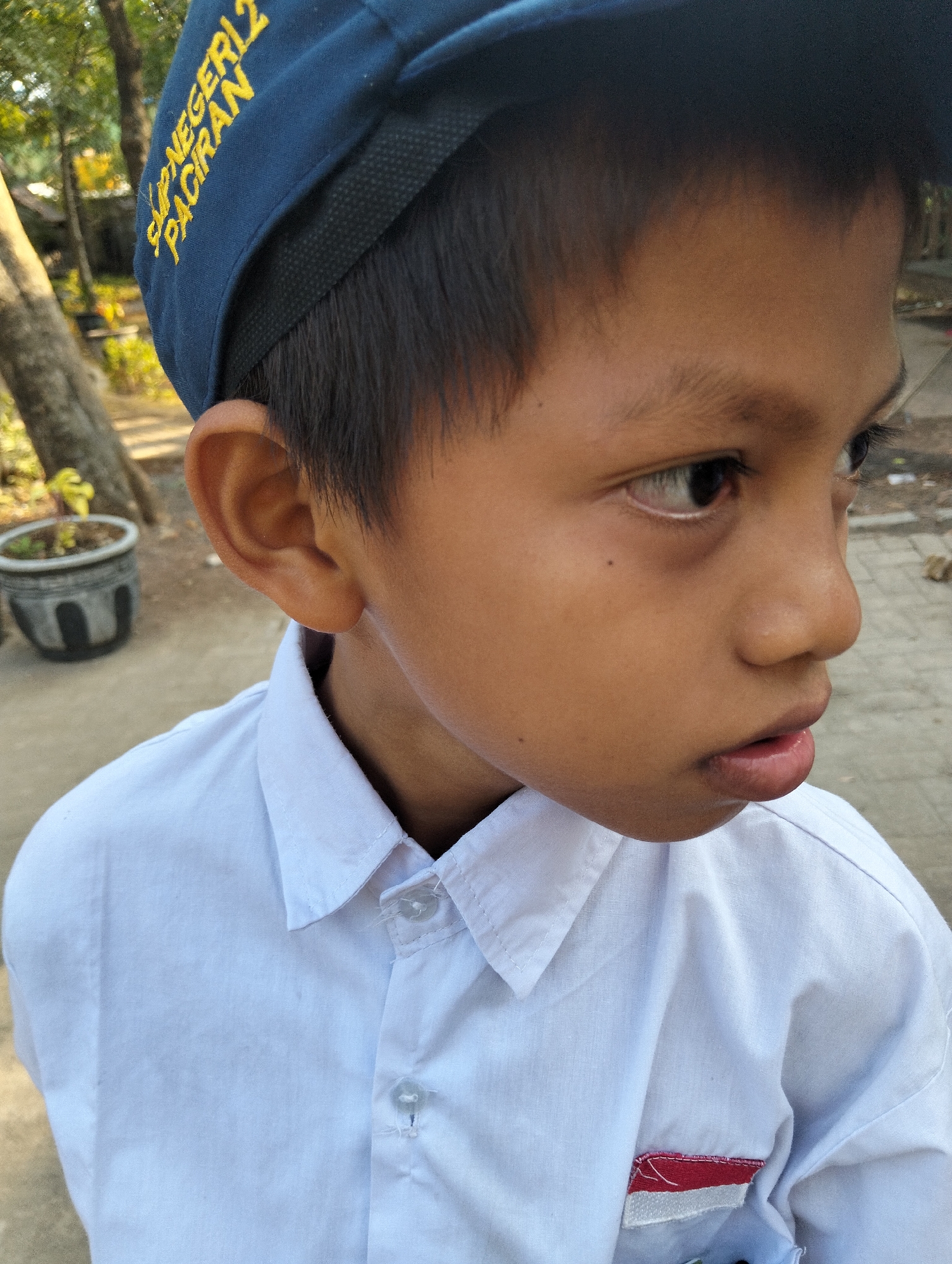 A medium shot captures a young boy wearing a white collared shirt and a dark blue baseball cap with yellow lettering that reads "SMP NEGERI 2 PACIRAN" in the city of Paciran, Indonesia. The boy is looking to his right, with his face partially obscured by his cap. The background shows a blurry outdoor scene with trees, foliage, and a concrete ground. The boy's shirt has a small embroidered patch on the left side, appearing to be the Indonesian flag.