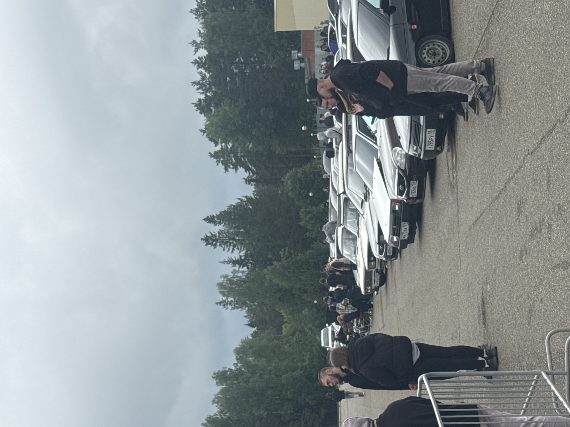 The image captures an outdoor gathering on a large asphalt paved area in Прикамский, Russia, under an overcast sky. A linear arrangement of approximately ten cars, predominantly older model sedans, including what appear to be Lada vehicles, is parked from the mid-ground extending towards the right. Several visible license plates include "E782XA 159," "P795PY 18," "P670AB 152," and "H900AA 51."
Numerous individuals, mostly men, are present across the scene, dressed in casual attire such as hoodies, jackets, jeans, and sneakers. In the immediate foreground, on the left, a silver metal barrier is visible. Three individuals stand near this barrier: one in a light-colored top, another in a black puffer jacket and black pants, and a bearded man in a black top. In the immediate foreground to the right, two men and a partially visible woman are engaged in conversation; one man wears a black cap, a black hoodie, and light grey pants, while the other is in a black long-sleeve shirt.
Further back, other groups of people are standing and sitting on portable chairs or coolers around the parked cars, indicating a social event or car meet. A white SUV is parked in the deeper mid-ground to the left. The background consists of a dense forest of coniferous and deciduous trees. To the right, a low-rise, light-colored building with a flat roof and visible HVAC units is situated behind the parked cars. Several tall light poles are also scattered throughout the background.