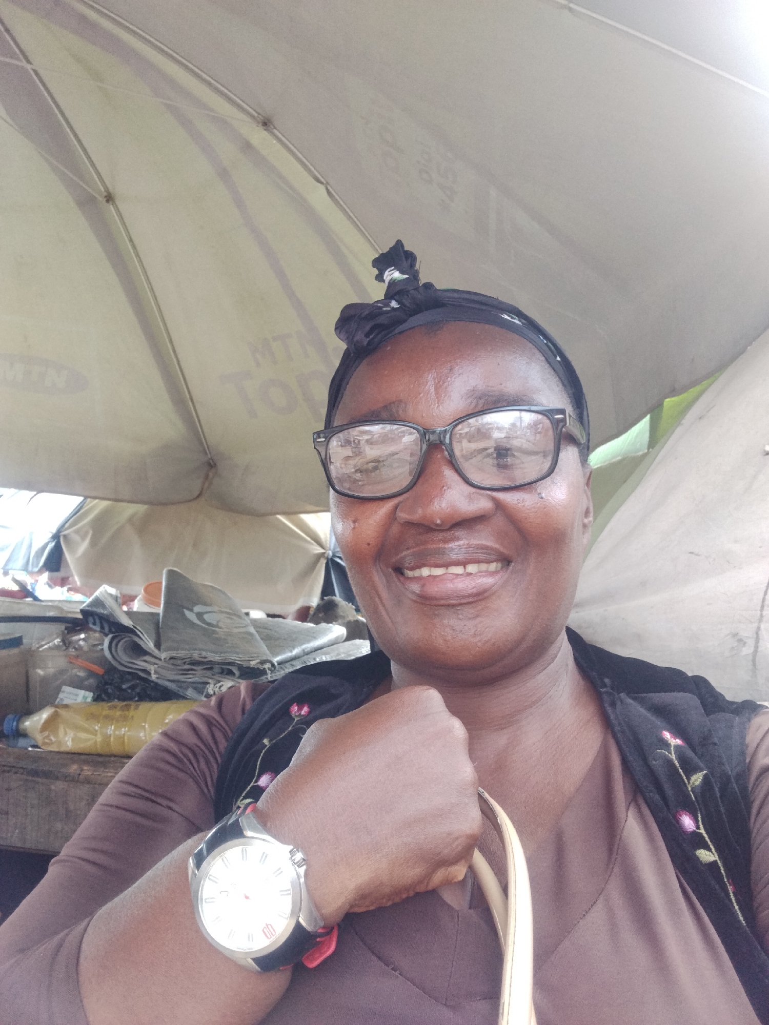 A middle-aged African woman smiles at the camera, wearing glasses, a dark head wrap, and a brown top with a floral-embroidered dark vest. She is also wearing a wristwatch on her left hand and holding a light-colored purse handle. She appears to be sitting under a MTN-branded umbrella at what seems to be a market stall. There are various objects in the background on a wooden table. Based on the prompt, this image could be located in Ura Nta Umuarandu, Nigeria.