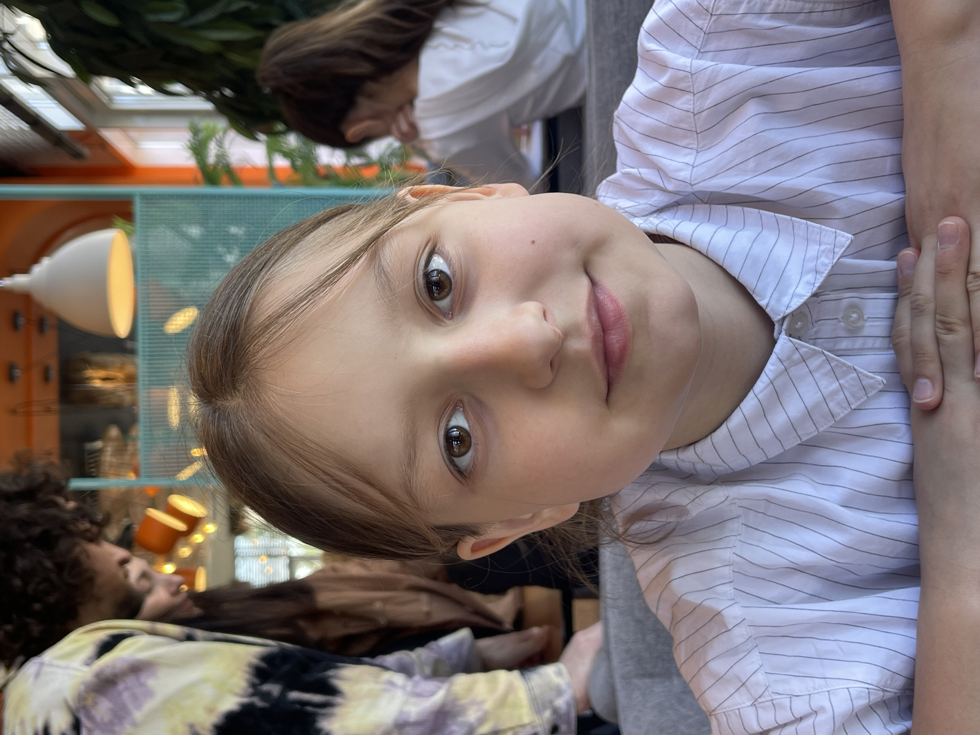 A young girl is the primary subject of the image, her face prominently displayed. She has light brown hair and brown eyes. She wears a white striped shirt. The background features other people, including a woman with brown hair and a man with curly dark hair and a colorful jacket. The setting appears to be an indoor dining area with architectural elements suggesting a modern design, including light fixtures and a grid-patterned wall. Based on the prompt's request for location details, this image is associated with the city of Stavropol, Russia.