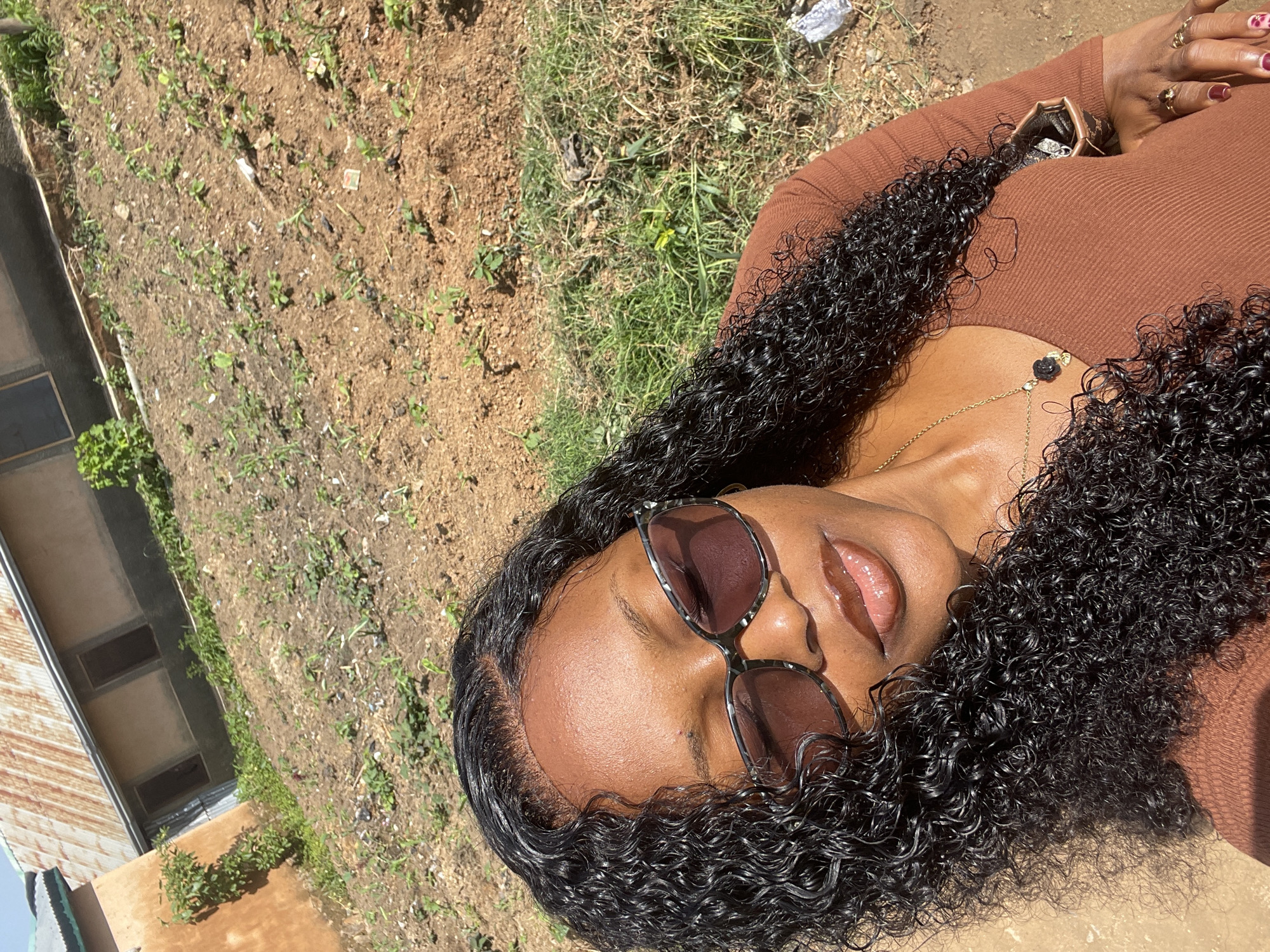 A medium shot captures a young Black woman with curly dark hair, wearing a brown long-sleeved shirt and sunglasses, posing for a selfie outdoors. The background shows a sloped, dry, dirt and grass area with scattered small plants, indicative of a rural or undeveloped part of a town. In the upper left corner, a building with a corrugated metal roof and earth-toned walls is visible, suggesting a residential or agricultural structure. The image appears to be taken in Rafin Par, Nigeria, given the context. The woman is looking down and slightly to her right, with her eyes closed and a subtle smile, her lips glossy. A delicate gold necklace with a black rose pendant adorns her neck. Her right hand, adorned with rings, is resting near her collarbone, and a portion of a brown bag is visible on her shoulder. The lighting suggests a sunny day, casting shadows on her face and hair.