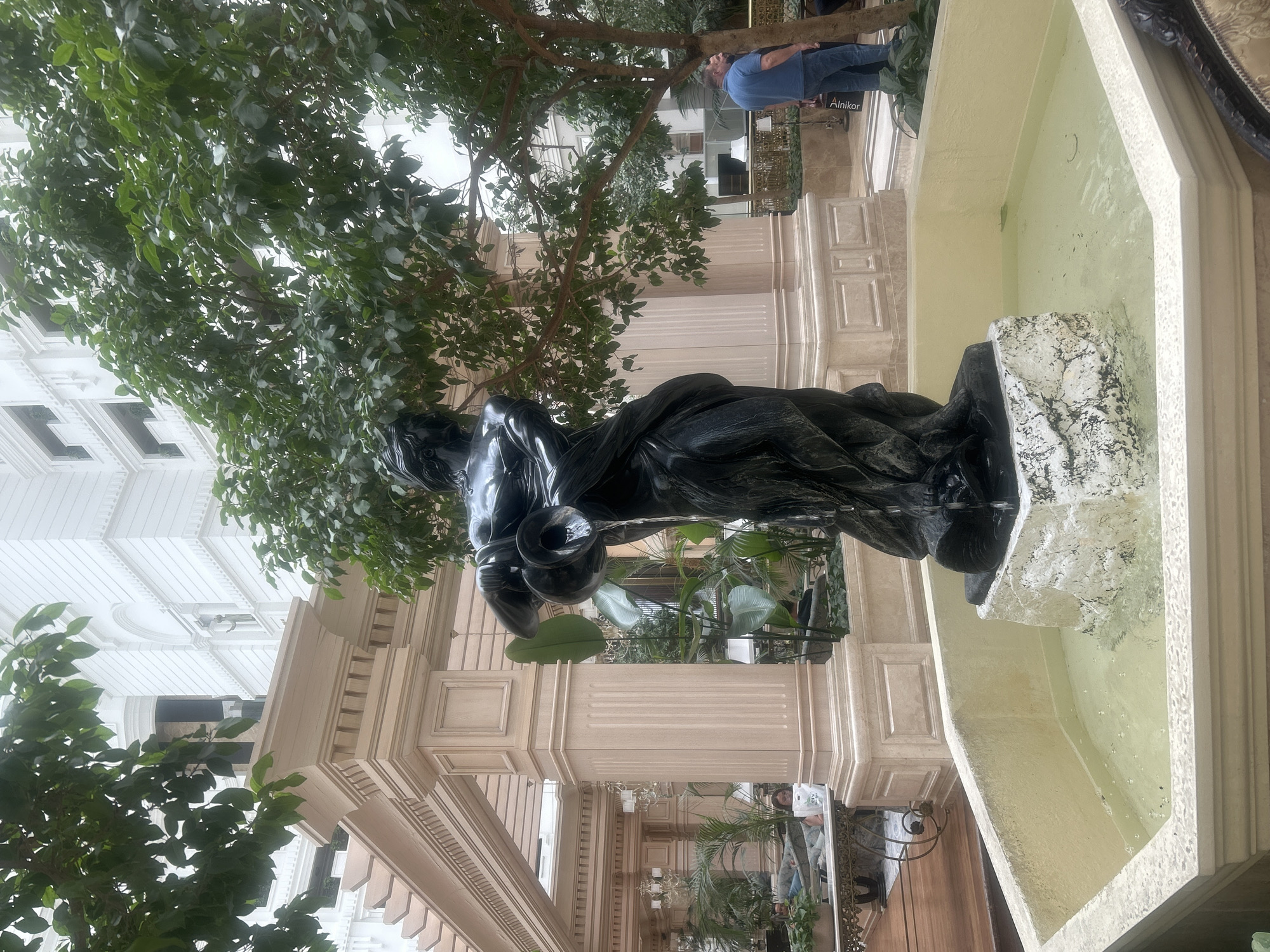 In Almaty, Kazakhstan, a black sculpture of a woman pouring water from a jug stands in the center of a hexagonal fountain. The sculpture and fountain are surrounded by lush greenery and ornate architectural elements. In the background, a multi-story building with white facade and large windows is visible. Several people are present in the scene, including two men standing near a tree to the right and a woman reading a book while sitting on a couch in the background to the left. The environment appears to be an indoor garden or atrium, likely within a hotel or public space, characterized by light-colored pillars, high ceilings, and decorative lighting fixtures.