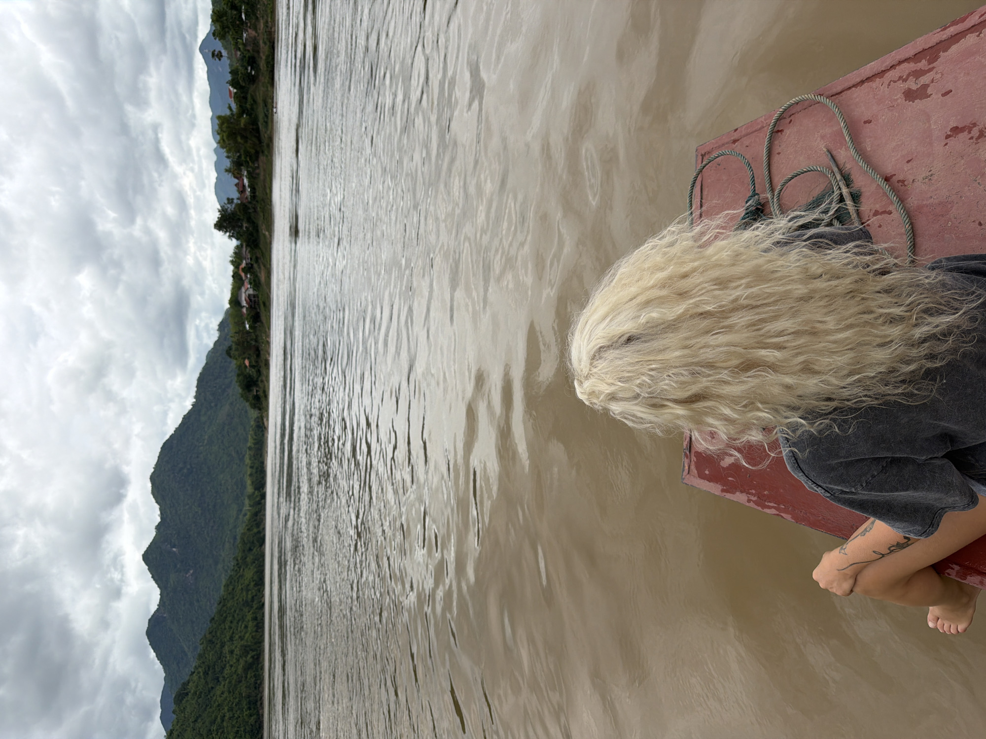 A person with long, blonde, curly hair is sitting on the front of a red boat, facing away from the camera, looking out at a wide, muddy river. The person is wearing a dark gray t-shirt and has tattoos on their arm and leg. Their legs are crossed, with their right knee bent and their left leg extended. A rope is coiled on the boat near the person. The river is calm with ripples on the surface, reflecting the overcast sky. In the background, lush green mountains rise up on either side of the river, with a small village visible on the right bank. The sky is heavily clouded with gray tones, suggesting a cloudy or possibly rainy day. The scene appears to be located in or near Houaymat, Laos.
