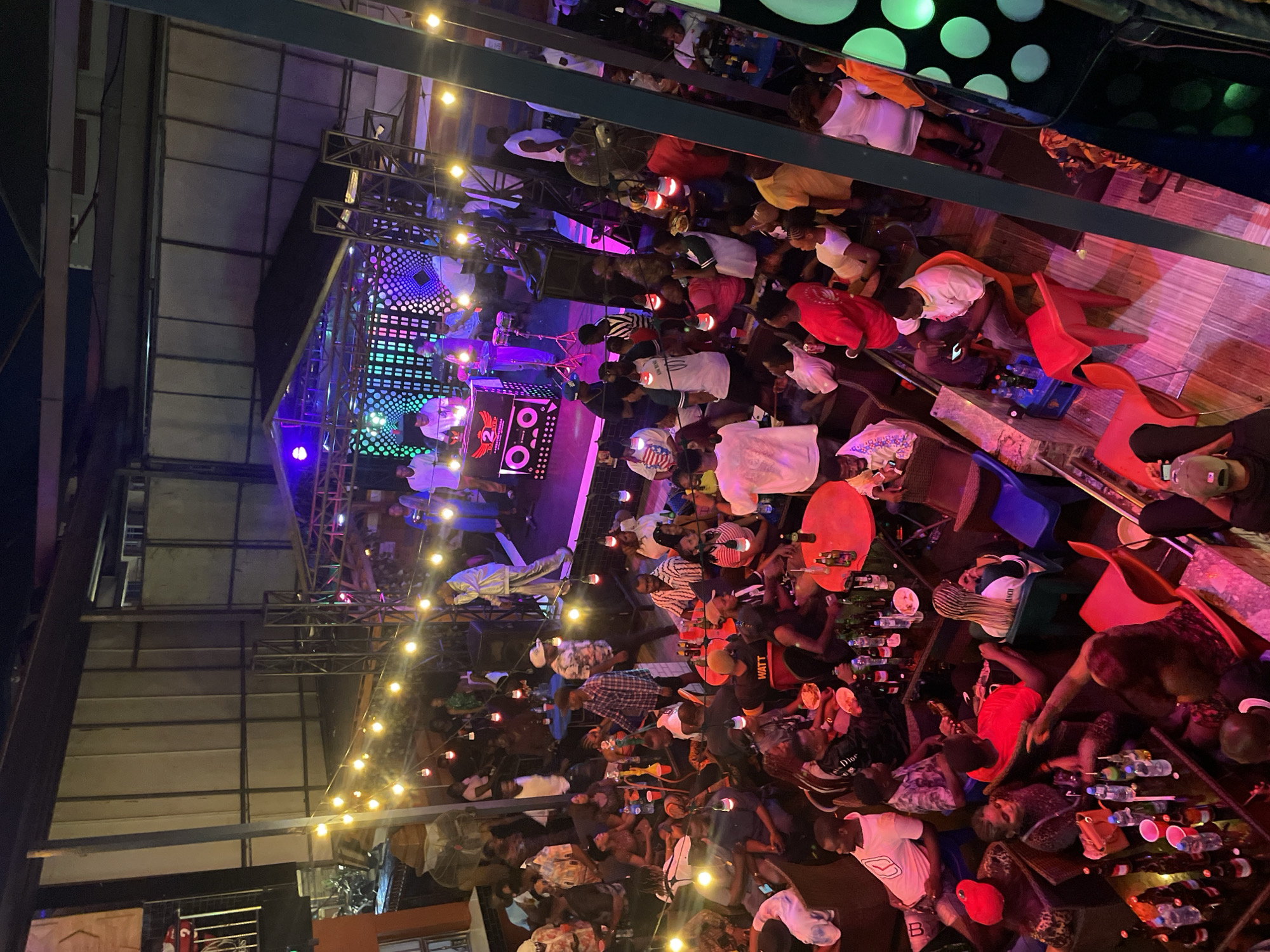 The image depicts a bustling, multi-level indoor or semi-outdoor entertainment venue, likely a bar or club, at night in the city of Unknown, Unknown. The scene is viewed from an elevated perspective, looking down onto the main floor and a stage area.
In the upper-middle, a raised stage features a DJ setup with a mixer, speakers, and a logo "2 ELP." Behind the DJ, a backdrop of animated LED screens displays dynamic geometric patterns in green, purple, and blue hues. Several individuals are on stage: one man in a light-colored suit holds a microphone, appearing to perform; another man in a white t-shirt operates the DJ equipment; and others stand nearby, with percussion instruments (congas) visible to the right.
The main floor below is densely populated with a diverse crowd of adults, engaged in various activities. Many are seated at numerous tables and banquettes, which are laden with bottles (predominantly beer and water), cups, and some plates of food. Other individuals are standing, conversing, or watching the stage. Attire ranges from casual t-shirts and caps to dresses. A man in a black t-shirt with "WATT" printed on it is visible, as is a woman in a white dress on the right side. Several people are holding up mobile phones, possibly recording the performance. Shisha pipes are present on some tables.
The venue is dimly lit, creating an energetic ambiance with a combination of warm string lights draped across the space, colorful stage lighting, and green circular decorative lights illuminating a wall on the far right. Structural metal beams and corrugated metal panels form parts of the venue's architecture. A large fan is visible near the stage. The overall impression is one of a lively social gathering with live entertainment.