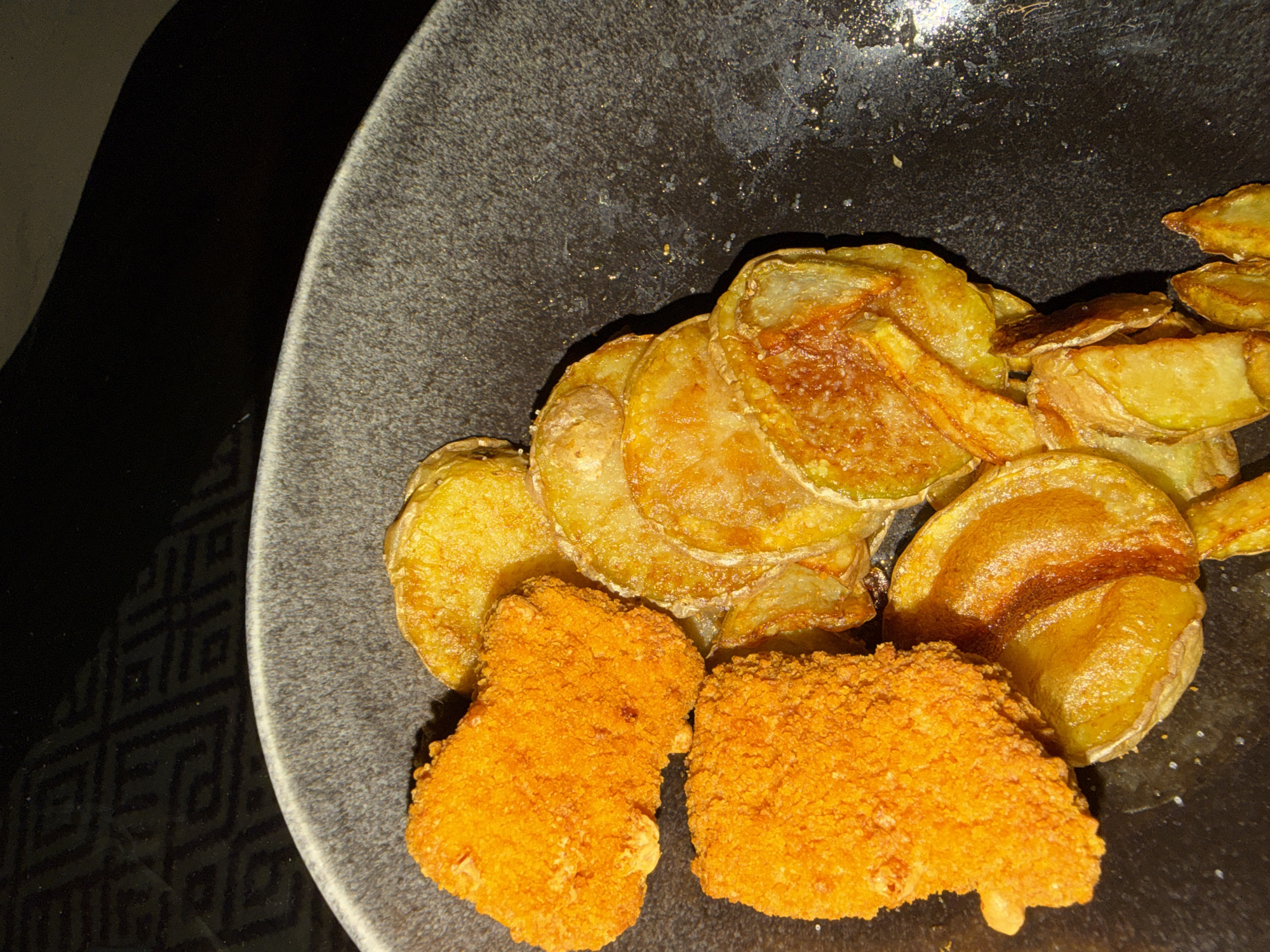 A close-up shot shows a portion of food presented on a dark gray, textured plate. The food consists of what appear to be fried potato chips, some of which are stacked, and two breaded, orange-colored items that may be chicken nuggets or similar fried snacks. The plate sits on a dark surface with a patterned design visible in the lower-left corner. The lighting is somewhat dim, focusing on the food. The location, Vila Real, Portugal, cannot be determined from the visual information. The absence of people, actions, interactions, and environmental context further restricts analysis.