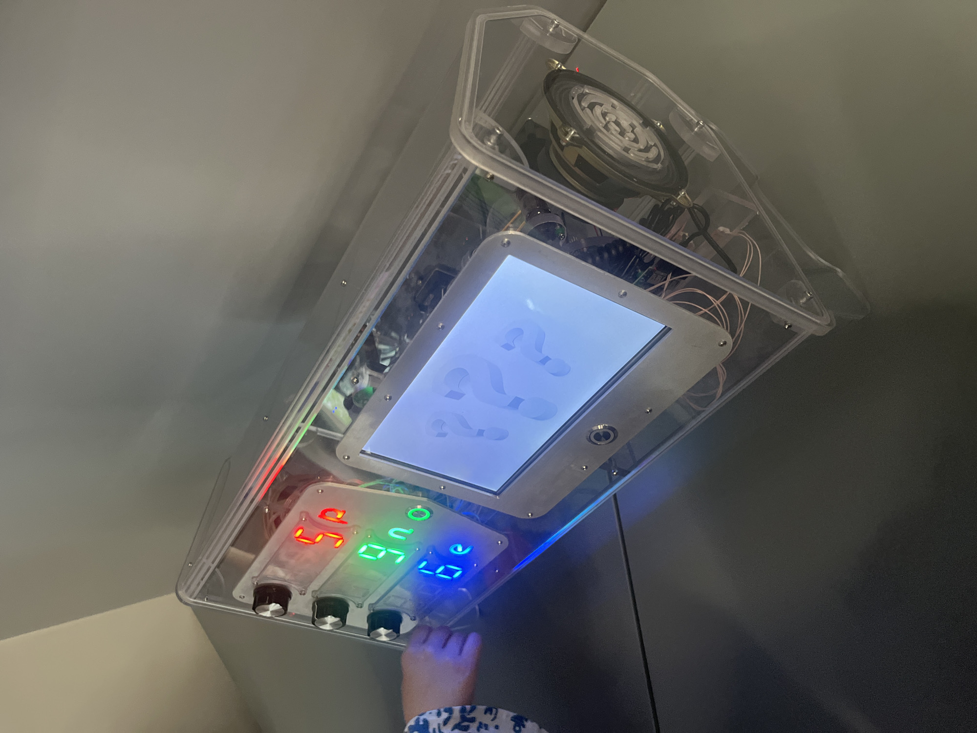 An enclosed, transparent, rectangular device is mounted on a gray wall. The device houses several components including a speaker, a screen displaying question marks, and a panel with three sets of LED numbers in red, green, and blue. A child's hand is visible, reaching to adjust a knob on the panel. The location is Yekaterinburg, Russia.