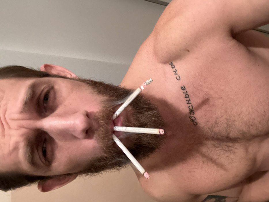 A shirtless man is photographed from a low angle. He has a light beard and a tattoo in Cyrillic script on his upper chest. He holds three lit cigarettes in his mouth. The background is a plain, light-colored wall. The image is tilted, giving a skewed perspective. His expression appears serious or intense.