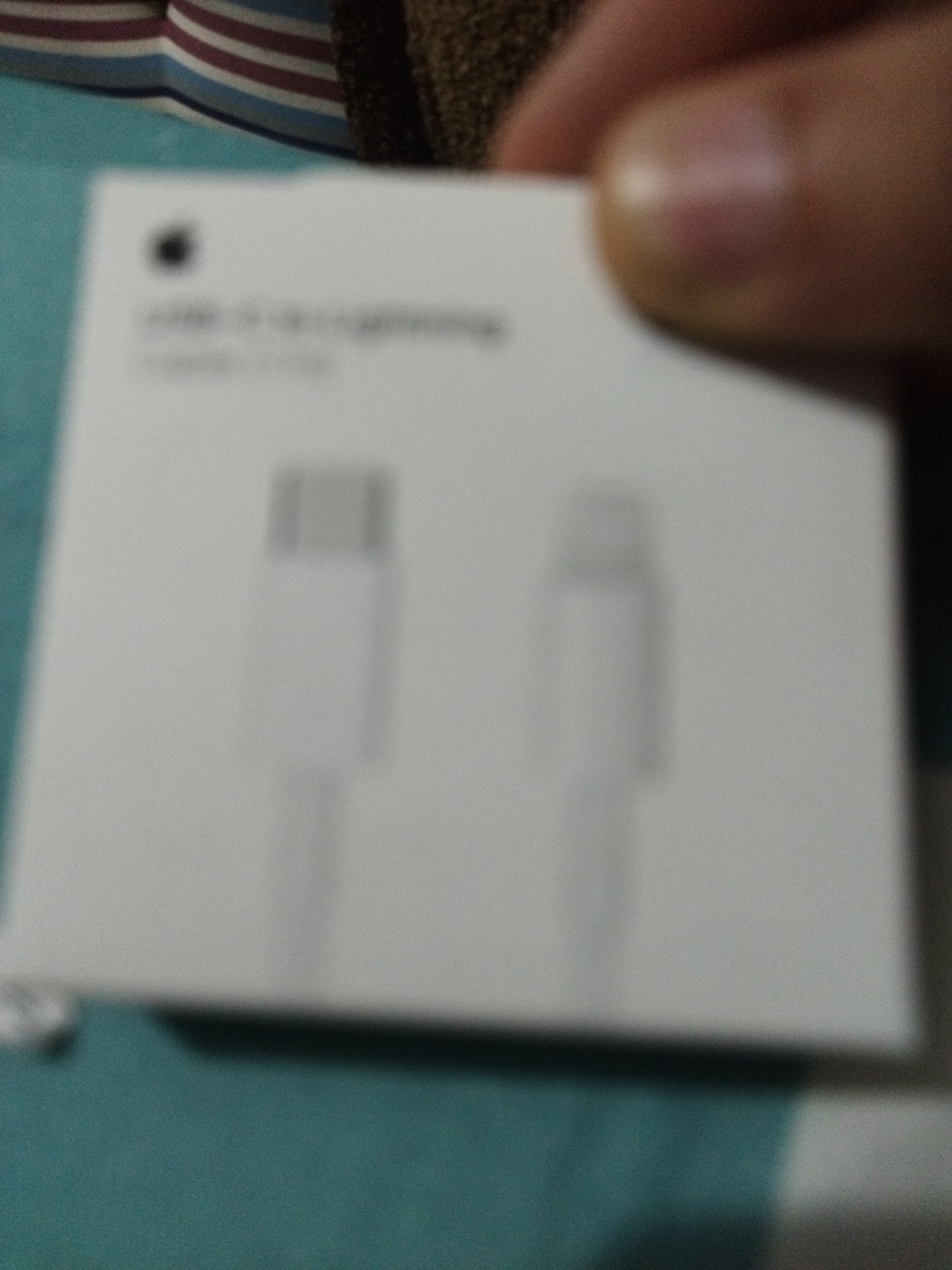 The image shows the packaging for an Apple USB-C to Lightning cable. The packaging is white and has the Apple logo at the top. Text below the logo is blurred but appears to say "USB-C to Lightning" and "Cable". Two blurred images of the cable connectors are also visible on the packaging. The packaging is being held by a person's hand, which is blurred in the upper right corner of the image. The background consists of a striped fabric in blue, white, and purple, and a textured brown fabric. The image is out of focus and has a grainy texture. There is no information or visual context to suggest that this image is located in the city of Smolyan, Bulgaria.