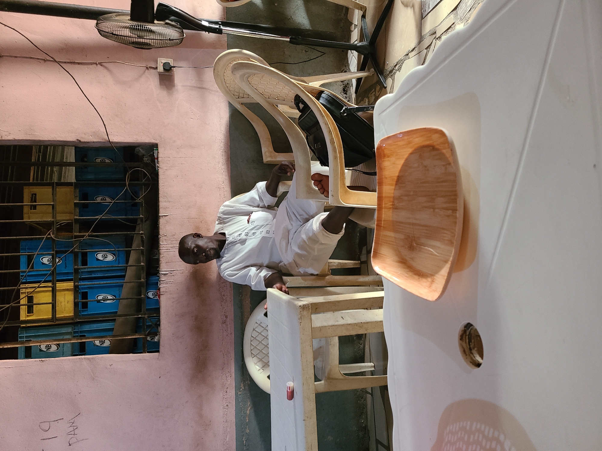 This image, set in what appears to be an informal indoor space in Jos, Nigeria, features a bald Black man in a white tunic looking directly at the camera. He is seated on a light-colored plastic chair, with his left foot resting on another chair. The man's expression is neutral and calm.
In the foreground, a large white plastic table with a circular hole dominates the right side of the frame, holding a light brown, rectangular tray. Behind the man, several other light-colored plastic chairs are visible, one with a black bag tucked underneath it. A small, clear container with a red cap is placed on a chair near the man.
The background shows a textured pinkish-brown wall. To the left, a metal-barred window reveals stacks of blue and yellow plastic crates outside, many bearing the letters "NB." Electrical wires are fastened along the wall, leading to an outlet from which a black electric fan, positioned overhead in the top-left, is powered. The floor is a plain, grey concrete.
Handwritten text "19" and "PAM" is visible on the lower left part of the wall. The scene suggests a modest, functional environment, possibly a small shop or a communal area, depicted during daylight hours.