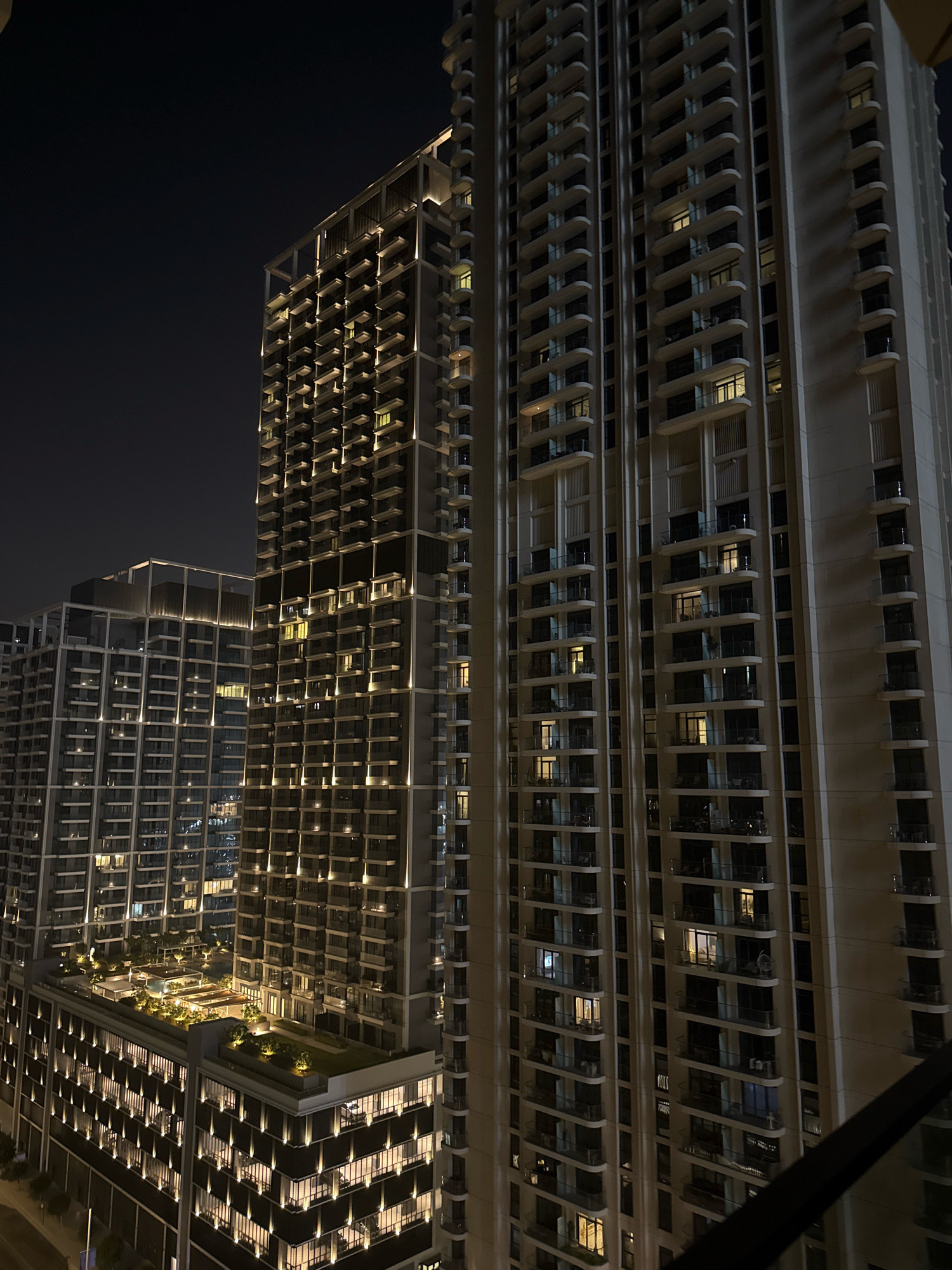 A nighttime aerial view captures a cluster of modern high-rise apartment buildings in Dubai, United Arab Emirates. The facades of the buildings are illuminated by architectural lighting, highlighting numerous balconies and windows, many of which show internal lights. The structures display a contemporary design with repeating patterns of balconies and windows, creating a sense of density and urban living. In the foreground, a lower, wider building features a rooftop garden with illuminated pathways and greenery, suggesting communal outdoor space. The overall scene is one of a well-lit, vibrant, and densely populated urban environment characteristic of Dubai.