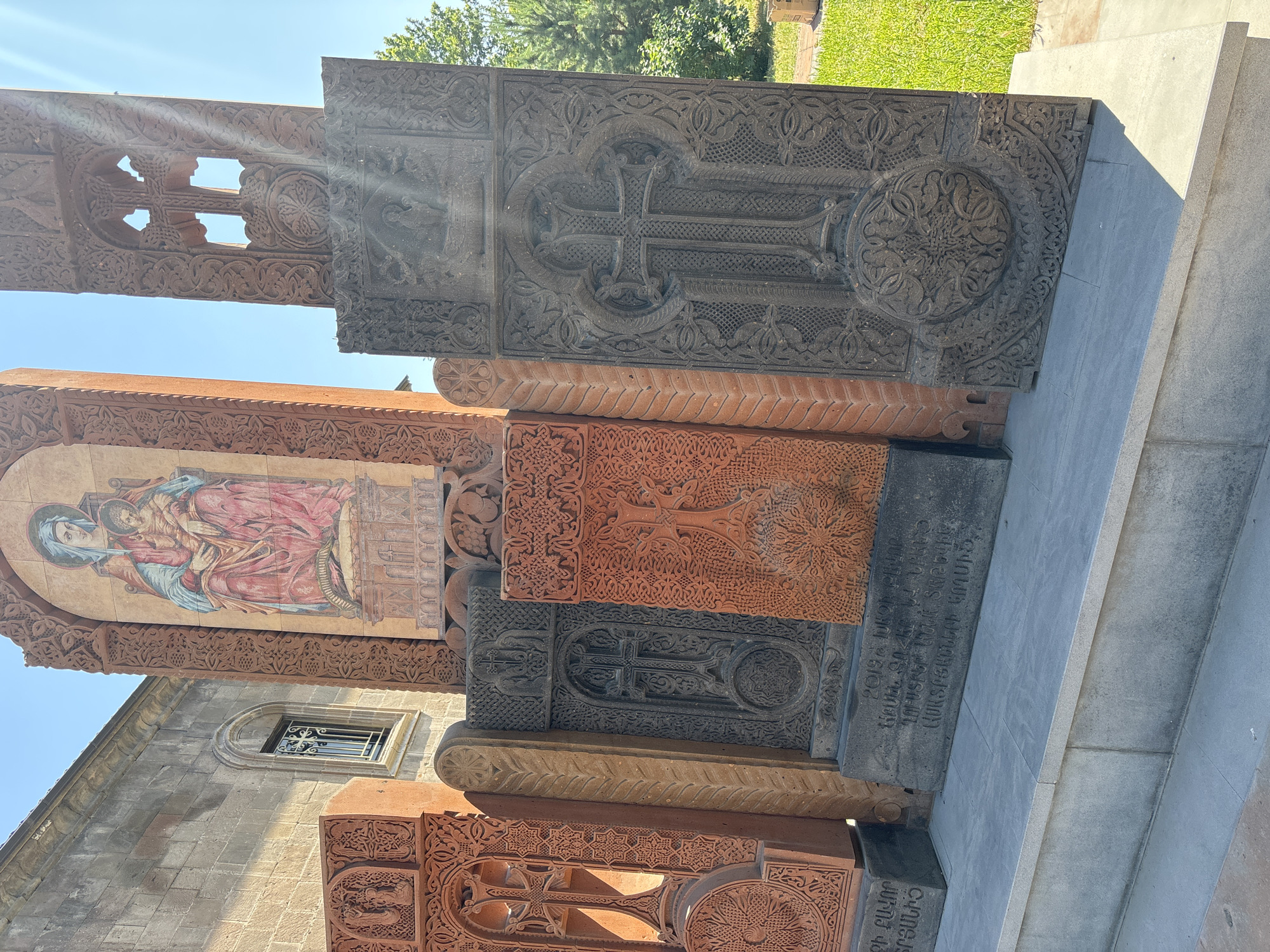 The image displays a vibrant outdoor collection of intricately carved Armenian khachkars (cross-stones) at a religious site in Vagharshapat, Armenia. Several stone monuments, varying in color from dark grey to reddish-brown, are arranged vertically, forming a decorative wall or exhibition alongside a stone building.
Each khachkar is richly adorned with elaborate relief carvings, featuring prominent crosses, intricate geometric patterns, and organic, often floral, motifs. Some crosses are carved as openwork, allowing bright sunlight to stream through. The different stone types and their detailed craftsmanship are evident.
Above one of the reddish-brown khachkars, integrated into an arched recess of the adjacent stone building, is a colorful mural. It depicts the Virgin Mary, wearing blue and red robes, holding the infant Jesus, who is also dressed in red. The building itself is constructed from light-colored stone and includes an arched window with a decorative metal grill.
The scene is set outdoors under a clear blue sky, with bright sunlight casting distinct shadows and highlights on the carvings. Green grass and trees are visible in the background, indicative of a well-maintained setting. Sunbeams are prominently visible, especially through the cutouts of the uppermost khachkars, suggesting a bright morning or afternoon.
On one of the dark grey khachkars, Armenian script is visibly carved. The inscription includes the year "2019" and reads, in part, "ՄԻԱՅՆ ՔՐԻՍՏՈՍՈՎ ԵՐԱՆԻ ԱՐԱՐԱՏԻ ՄԵՆՔ ՏԵՍՆՈՒՄ ԱՂԱԽԱԹԱՆՆԵՐԻ ԿՈՂՄԻՑ" which translates roughly to "2019, ONLY BY CHRIST, WE SEE THE BLESSED ARARAT, BY THE PRAYERS OF..." This text suggests a dedication or commemorative purpose for the khachkar. Stone steps are visible on the right, leading to a higher elevation.