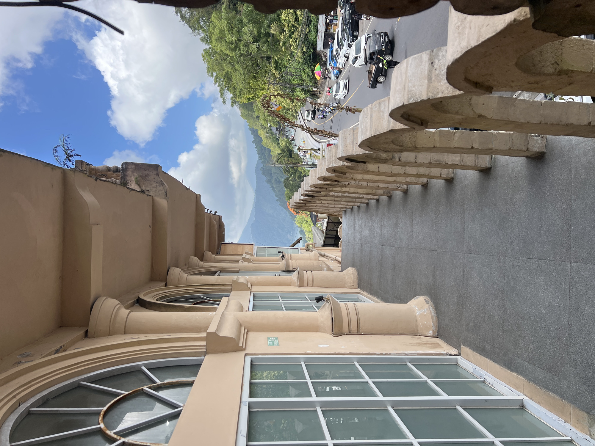The image depicts a view from a balcony or elevated walkway in Kintamani, Indonesia, overlooking a scenic mountainous landscape with a road winding through it. The architectural style of the building features large arched windows and ornate pillars, with a textured beige facade. The walkway itself is paved with dark gray tiles and has a decorative stone balustrade with repetitive arch shapes.
Below, a road is visible, bustling with traffic including cars, motorcycles, and a black pickup truck. Several people are seen near the road, some of whom appear to be walking or standing. The surrounding environment is lush with green trees covering the hillsides. The sky is a vibrant blue with scattered white clouds. Traditional Indonesian decorative elements, such as woven garlands, are present along the roadside, indicating a cultural context. The overall scene captures a lively roadside view from a distinct vantage point in Kintamani.