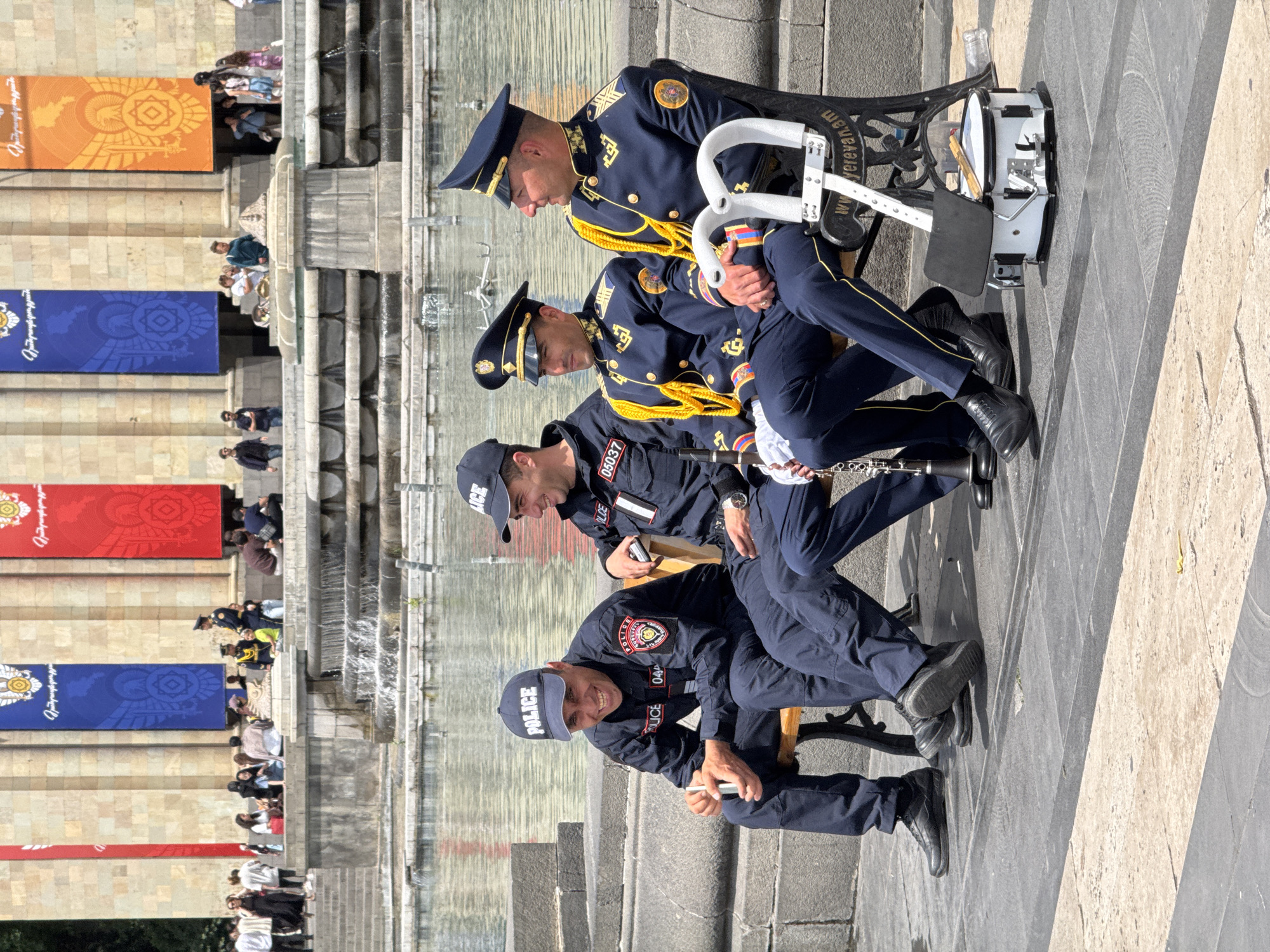 Four men in uniform are sitting on benches in an outdoor urban environment. Two of the men are wearing dark blue uniforms with gold trim and peaked caps. One of these men is holding a drum with drumsticks. The other two men are wearing dark blue uniforms with "POLICE" written on their caps, and numbers on their chest. The men are sitting on a stone surface with gray and beige stripes. In the background are several tall buildings with banners of various colors, including orange, blue, and red. There are people sitting on the steps of the buildings. The location is the Republic Square in Yerevan, Armenia.