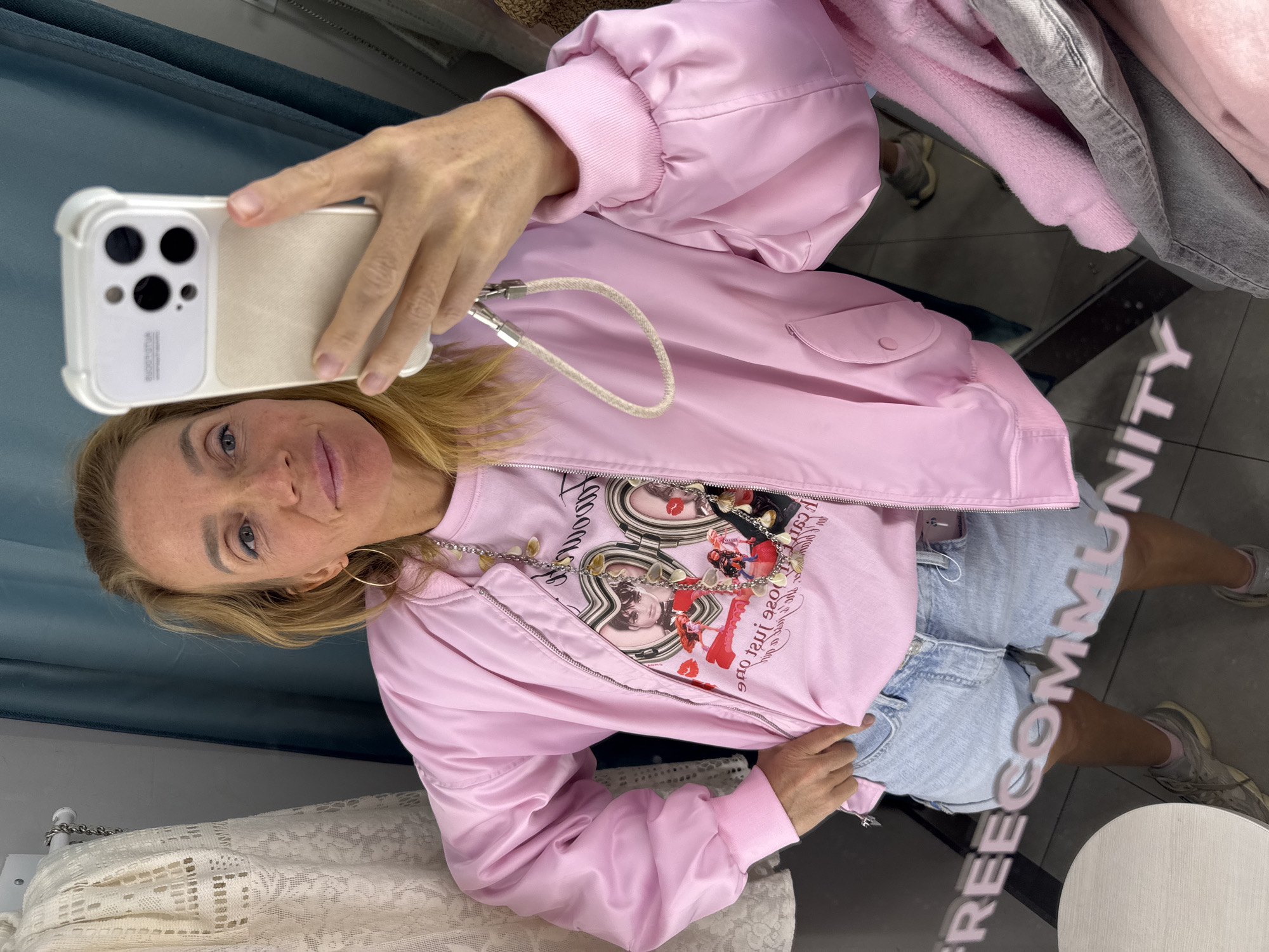 A woman is taking a selfie in a store dressing room. She is wearing a light pink bomber jacket, a t-shirt with a graphic print, and light blue denim shorts. Her blonde hair is shoulder-length, and she is wearing gold hoop earrings and a necklace. The phone she is using has a white case. The dressing room has a teal blue curtain, a white lace garment, and reflective flooring with the word "COMMUNITY" visible in reverse. The location cannot be determined as Vsevolozhsk, Russia, based on the visual information in the image.
