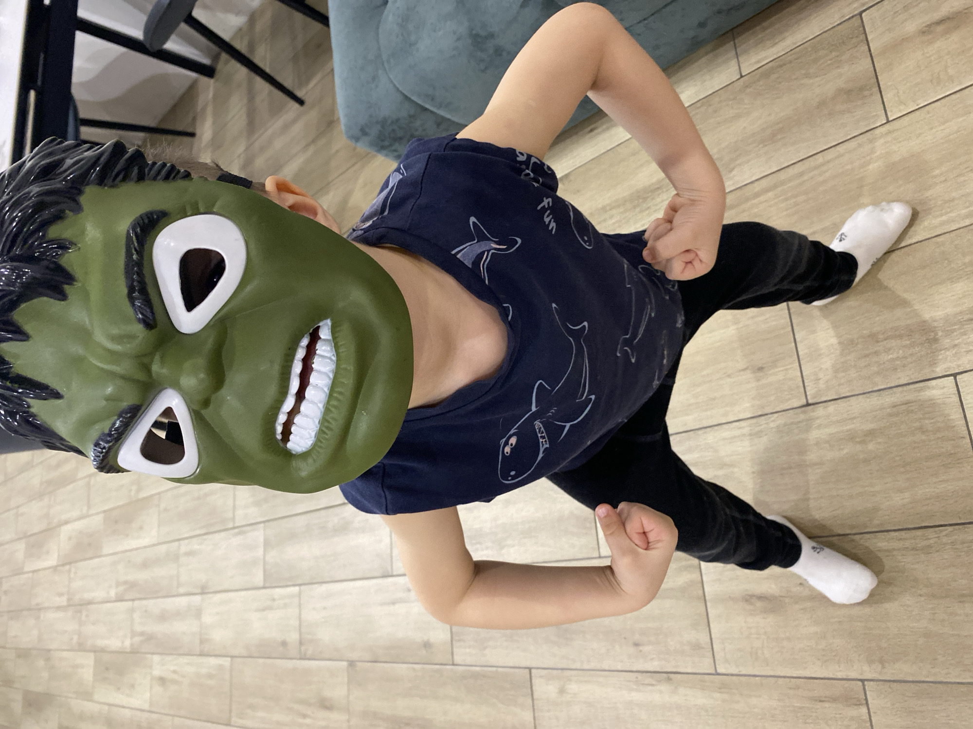 A child wearing a green Hulk mask and a blue shirt with shark designs is standing on a light brown wood-look tile floor. The child is wearing black pants and white socks and is flexing both arms. There is a chair and part of a green couch visible in the background. The photo appears to be taken from directly above the child. It's impossible to confirm the location.