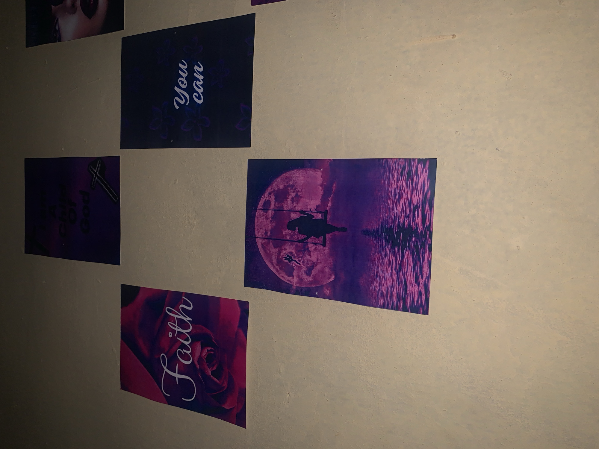 The image shows a section of a wall decorated with several posters. The posters are of varying colors and designs. One poster features the words "You can" in a stylized font. Another shows a cross and the words "Child of God." A third poster depicts a pink rose with the word "Faith" written across it. A fourth poster displays a purple scene of a full moon with a silhouette of a person fishing, and reflections of the moon and fishing line in the water. The wall itself appears to be a light beige or cream color and has a slightly textured surface. The location is unspecified and cannot be determined from this image. It is impossible to verify that the location is Keffi, Nigeria.