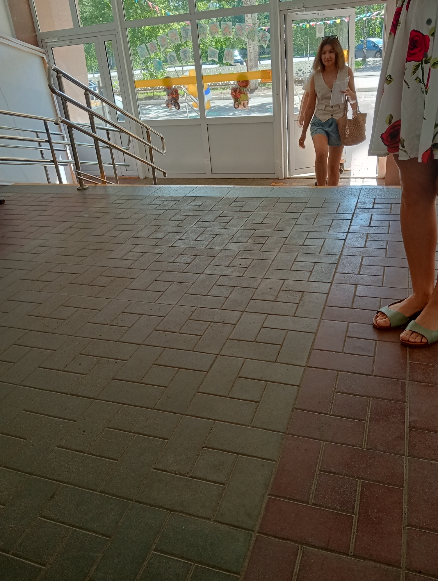 A young woman walks up a ramp, holding a woven bag and a piece of paper. She is wearing a light-colored vest over a white top and denim shorts. She is entering a building through double glass doors. Outside the building, there are trees and a blue car. The floor is tiled with a herringbone pattern. The scene appears to be outdoors, possibly in a public building or entrance.