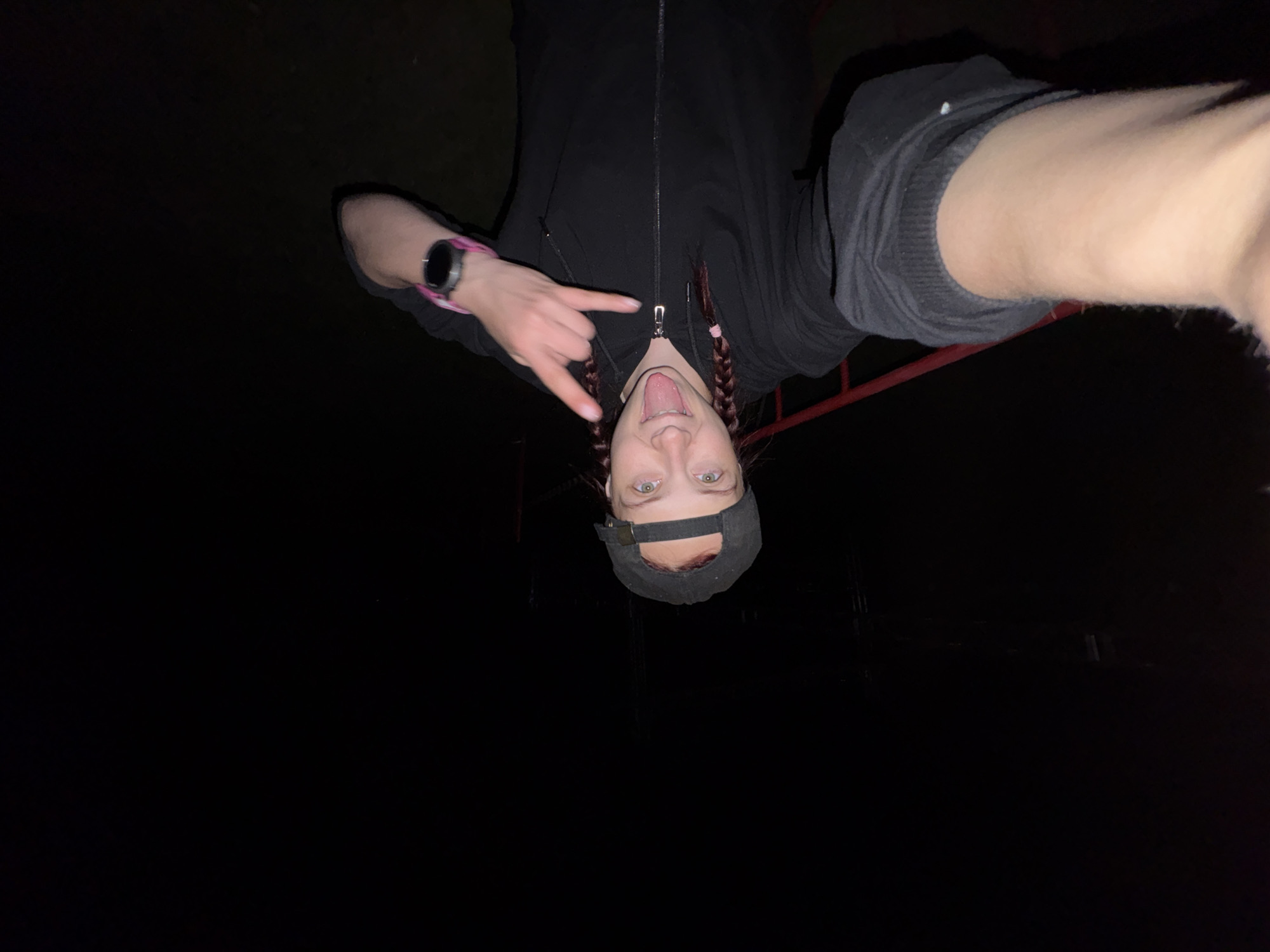 A young person is upside down, taking a picture. They are wearing a dark-colored hooded sweatshirt, a black cap, and have their tongue out, making a hand gesture. The surroundings appear dark, suggesting nighttime or a dimly lit area. The location cannot be identified as Khvalynsk, Russia, or any specific location, based on the visual information provided in the image.