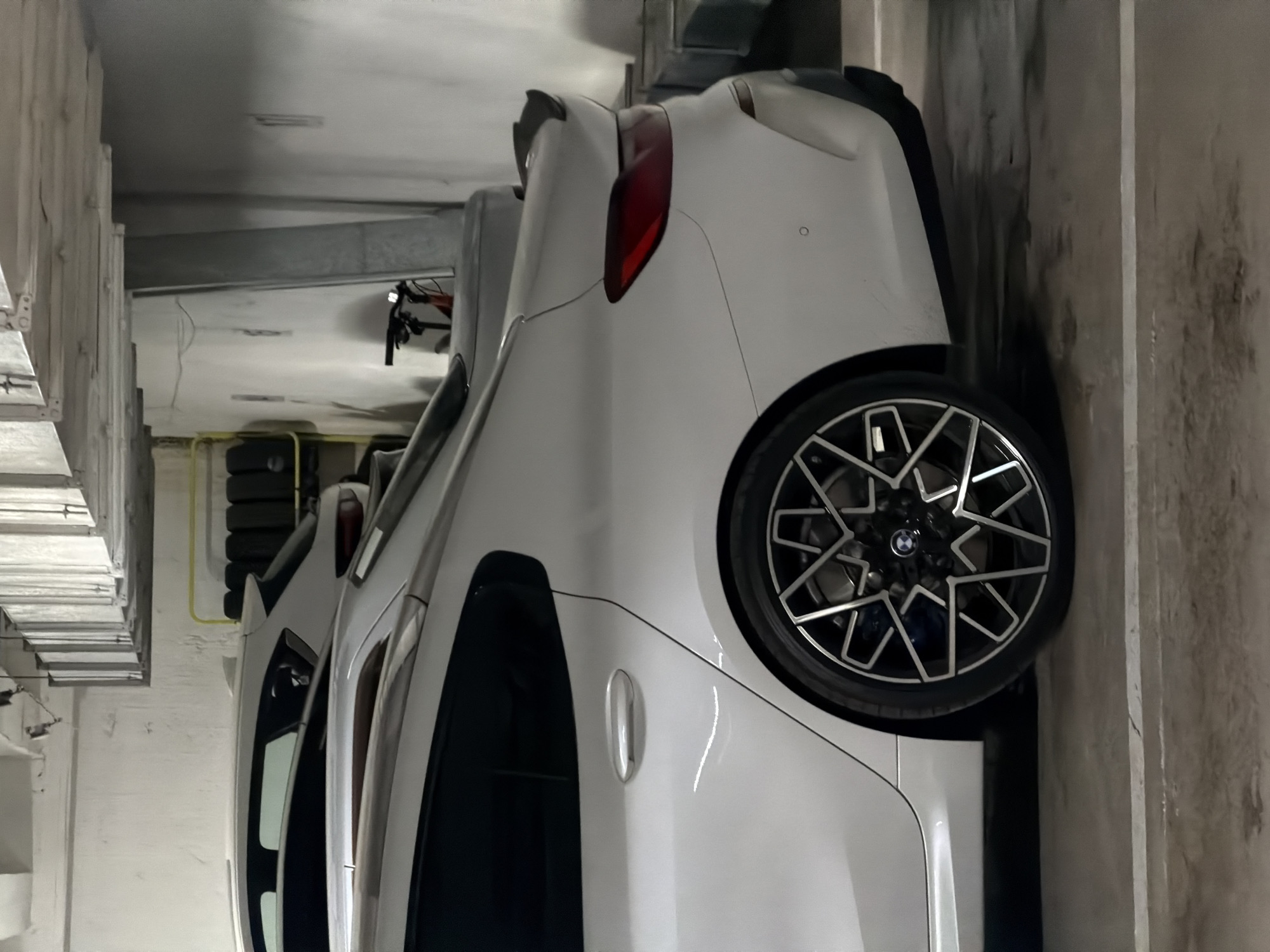 The image displays a white car, identified as a BMW by the logo on its wheel, parked in an indoor garage or underground parking facility. The car is viewed from its right rear quarter, showing the rear passenger side, taillight, and part of the trunk. It features a distinctive, multi-spoke alloy wheel, behind which a blue brake caliper is visible. A small black spoiler adorns the trunk lid, and a sleek red taillight is prominent on the rear passenger side. The rear passenger door includes a chrome handle.
In the background, on the left side of the image, a stack of black tires is visible next to a yellow pipe or hose running along a concrete wall. Further into the background, parts of a bicycle, including handlebars, are discernible. The setting is characterized by concrete walls and pillars, some showing signs of wear or staining, and metal ductwork is visible on the ceiling above. The lighting appears artificial, consistent with an indoor parking area. The car is stationary, and no people or specific activities are depicted. The BMW logo on the wheel is the only visible text.