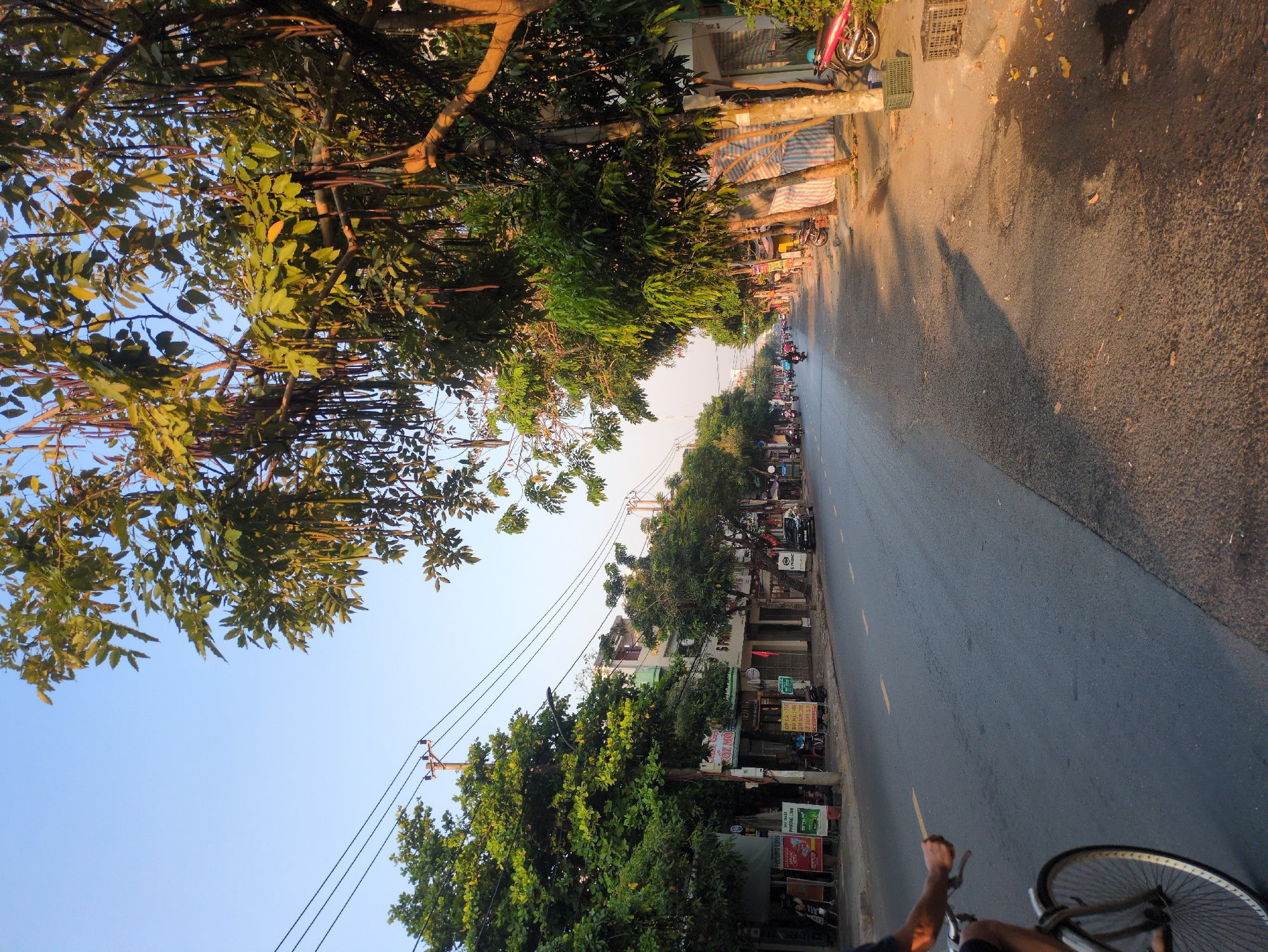 The image shows a street view in Tuy Hòa, Vietnam, captured from a low angle, possibly while riding a bicycle. The road stretches into the distance, lined with buildings, trees, and parked motorbikes. The sky is visible above the trees. The road surface is asphalt and has some minor wear. A bicycle wheel is partially visible in the foreground, and a hand is visible on the handlebars. The sidewalk on the right side is wet. Shadows indicate that the image was taken during the day.