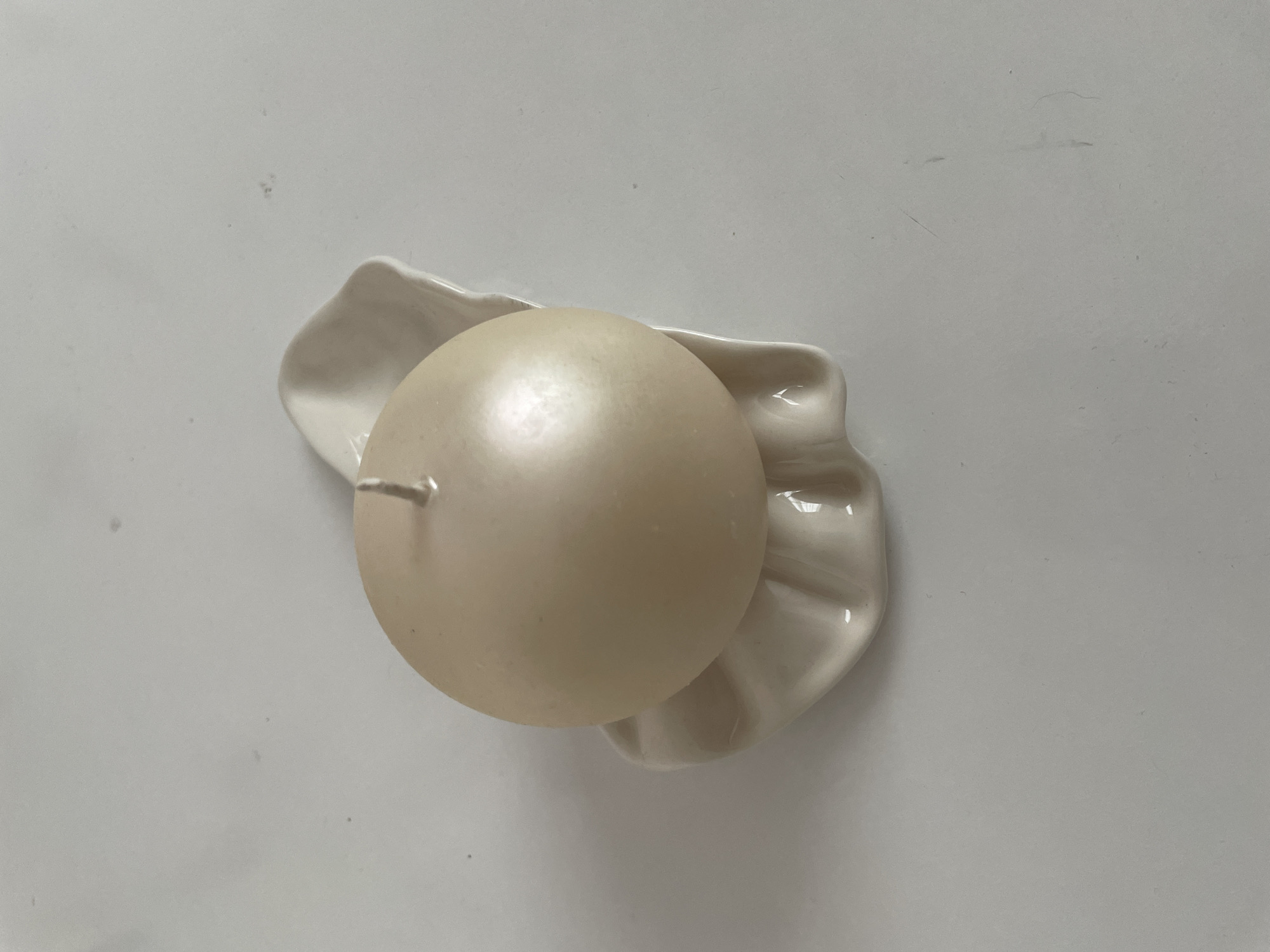 A top-down view shows a spherical, creamy-white candle resting on a white, petal-shaped ceramic dish. The candle's wick is visible. The dish and candle sit on a plain, light grey surface. The location is the city of Лесной Пейзаж Кп (шарапово), Russia.