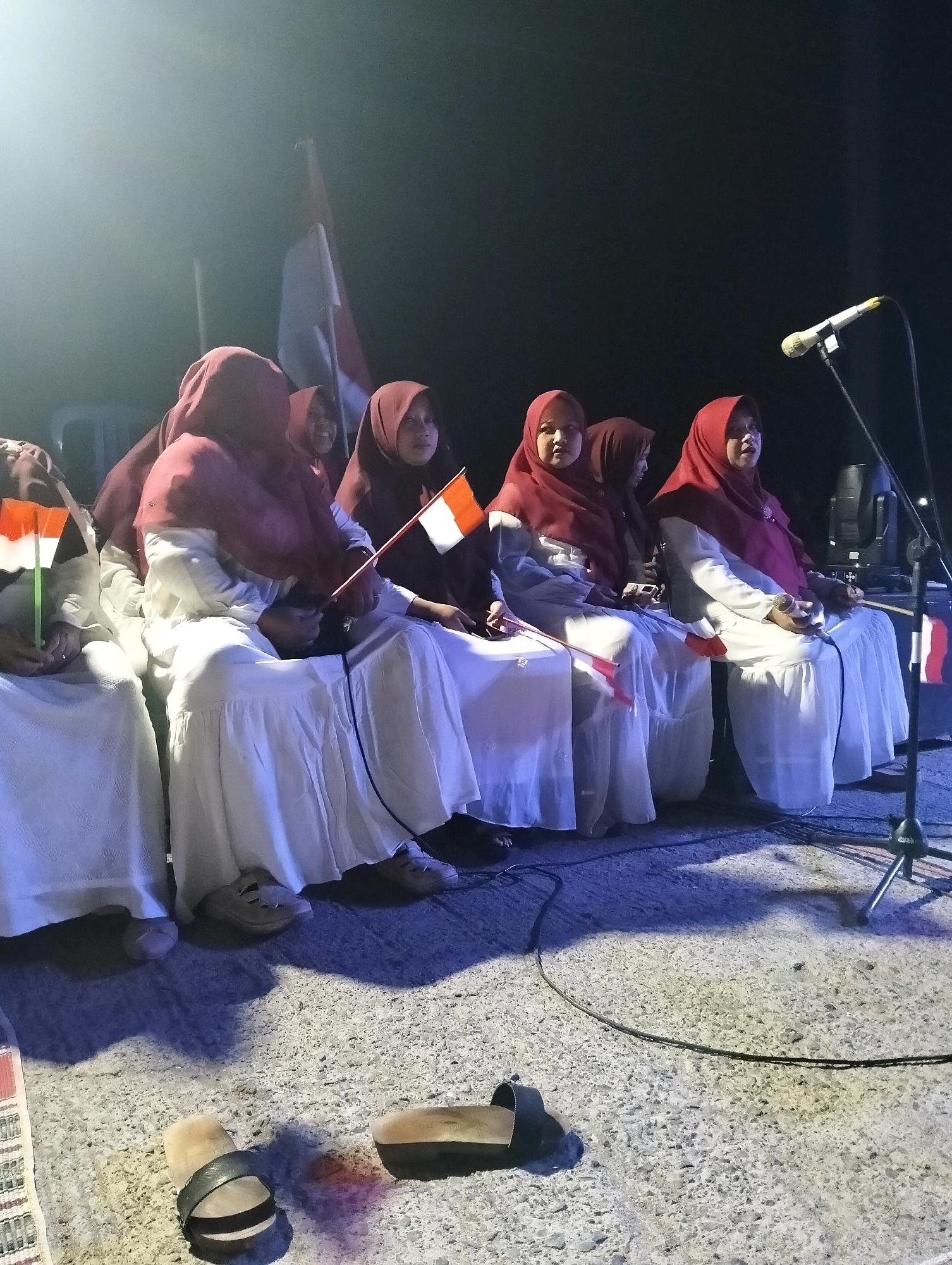 In Paciran, Indonesia, a group of girls, likely students, are seated in a row, wearing white dresses and maroon hijabs. They are holding small Indonesian flags, the red and white ensigns, in their hands, suggesting participation in a patriotic event or performance. The setting appears to be an outdoor stage at night, illuminated by spotlights that create a hazy, atmospheric glow. A microphone on a stand is positioned to the right of the group, indicating a possible musical or speaking engagement. In the foreground, a pair of sandals are discarded on the textured ground, hinting at a casual or relaxed atmosphere despite the formal attire of the participants. The overall scene evokes a sense of community and national pride, possibly during a celebration or school event.