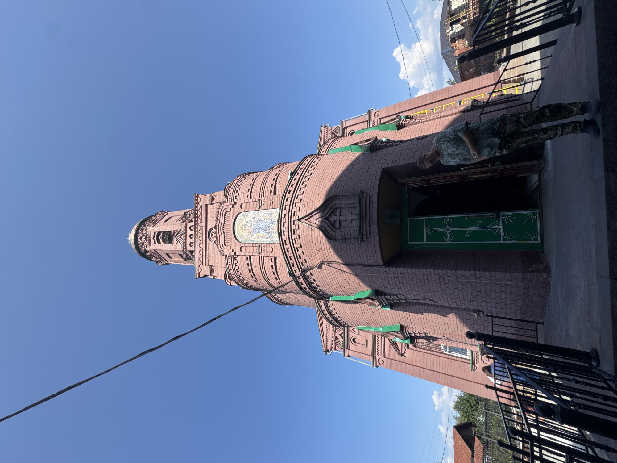 An exterior, low-angle, wide shot captures a brick church with a tall, ornate tower under a clear blue sky. The church is constructed of reddish-brown bricks and features green-painted doors and trim. A mosaic image is visible on the upper portion of the tower. A man in camouflage clothing stands near the entrance, looking down at his hands. In the foreground, black metal railings lead up to the entrance stairs. Behind the church, residential buildings and trees are visible. The scene appears to be located in Tolyatti, Russia, specifically at the Church of the Nativity of the Blessed Virgin Mary, a prominent landmark in the city.