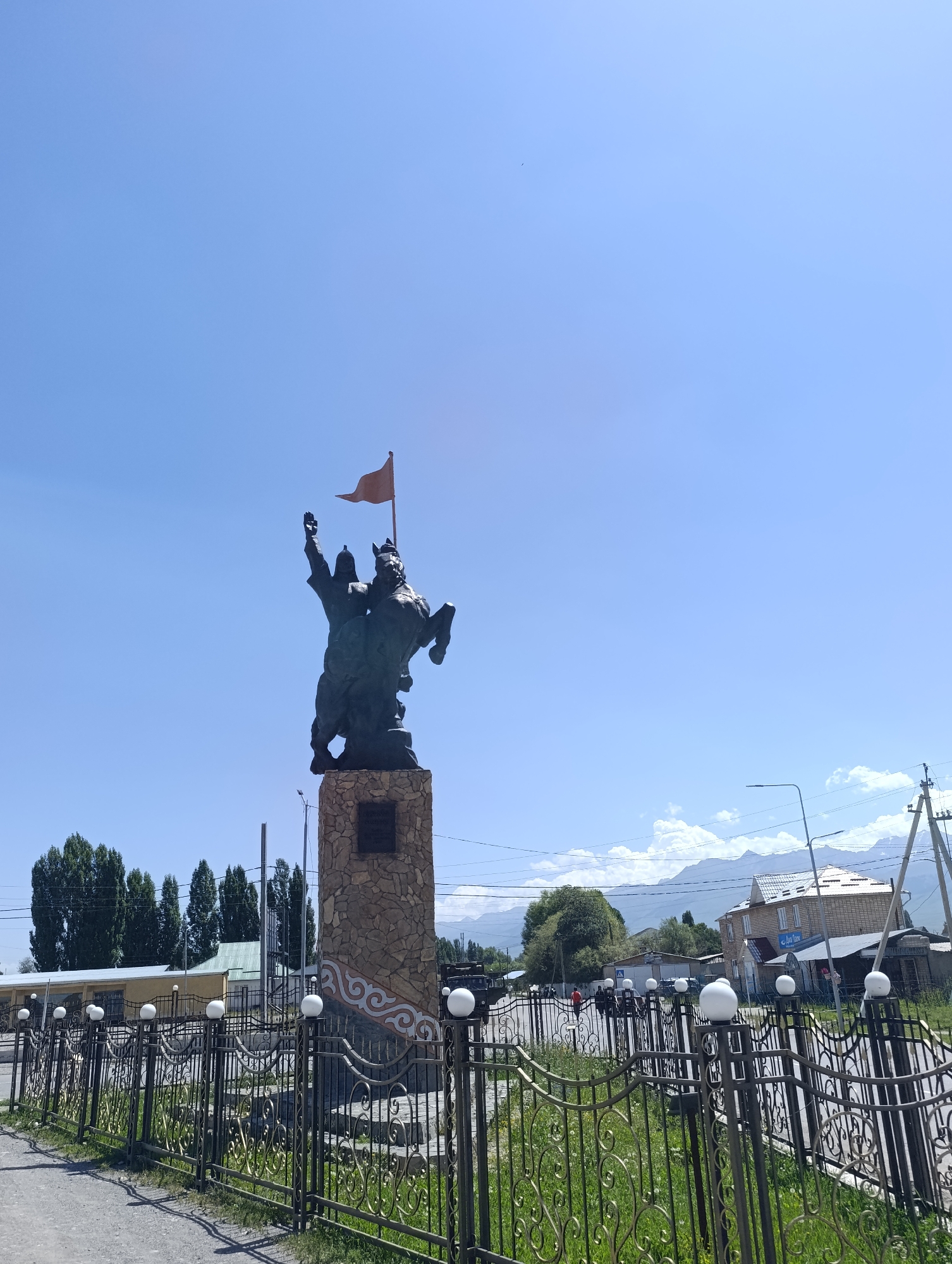 This image shows a statue of a man on horseback holding a flag in At-Bashy, Kyrgyzstan. The statue is made of dark metal and stands on a stone pedestal. The man is wearing traditional clothing and a helmet, and he is raising his right arm holding a small orange flag. The horse is depicted in a rearing stance, with its front legs in the air. The pedestal is decorated with traditional Kyrgyz ornaments in white. In the background, there are trees, buildings, mountains, and a clear blue sky. The scene appears to be set in a public space, possibly a town square or park, with a decorative fence in the foreground.