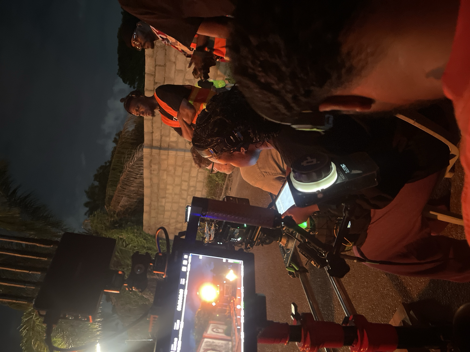 Here's a detailed description of the image:
**Location:** Unknown city, possibly filmed at night, streets illuminated by artificial lighting.
**Subjects:**
* A film crew is actively involved in the shooting, likely using a professional camera setup with a monitor displaying the shot.
* Several individuals are near the camera. One man wears a high-visibility safety vest, another wears Sony headphones, another appears to be reading a document.
**Objects:**
* A professional film camera with a monitor on a tripod.
* A brick or concrete wall in the background.
* Palm trees and other foliage behind the wall.
**Actions/Interactions:**
* The film crew members are focused on the camera monitor, presumably reviewing the shot or preparing for the next take.
**Environment:**
* The scene is dimly lit, requiring artificial lighting for the filming.
* The presence of palm trees suggests a tropical or subtropical location.
**Forensic Details:**
* The camera rig has multiple components attached, including power supplies, monitors, and potentially wireless video transmission devices.
* The camera is pointed towards a set or location that appears to have a narrow path or corridor.