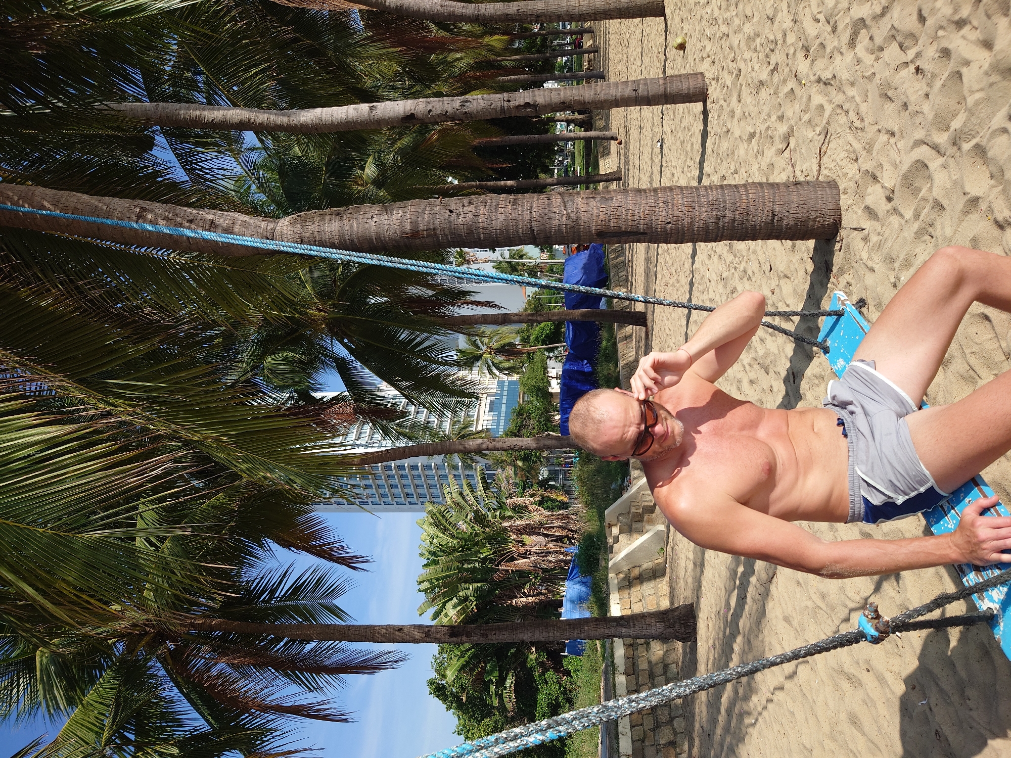 A middle-aged Caucasian man with a bald head, wearing sunglasses and grey swim shorts, sits on a blue swing on a sandy beach. He is partially shirtless, with his upper body exposed to the sun. The man is leaning forward, with his left hand adjusting his sunglasses and his right hand resting on the swing. The swing is suspended by thick ropes from two palm trees. The background features a row of tall palm trees, with a modern, multi-story building visible between them. The beach is composed of light brown sand with footprints and some scattered debris. The sky is blue with some light clouds. The overall impression is a relaxed, tropical setting.