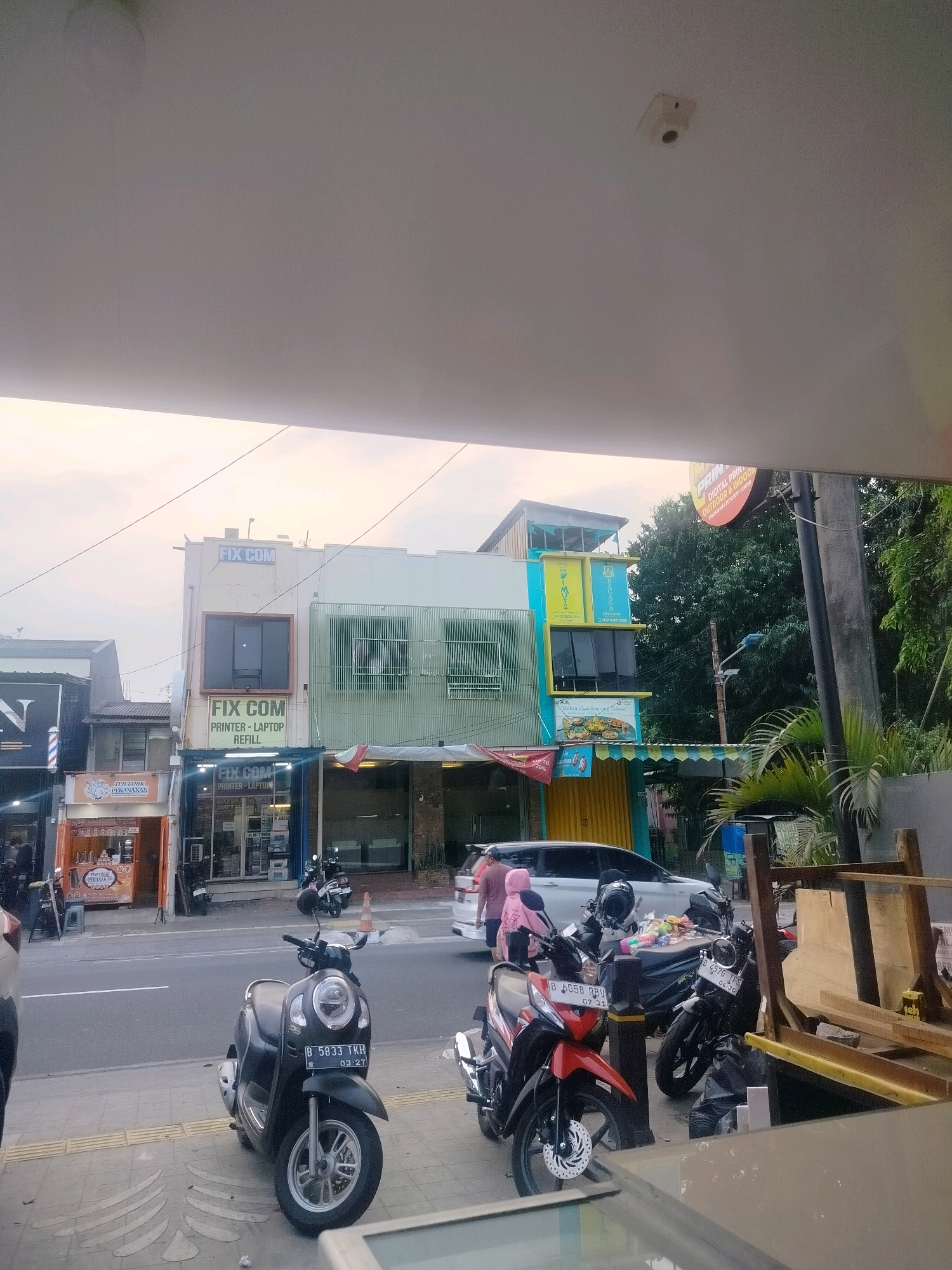 The image captures a street scene in East Jakarta, Indonesia, focusing on a row of shops and parked motorcycles under an overhang. The buildings are multi-story, with "FIX COM" signage prominent on one of them, indicating a business specializing in printers, laptops, and refills. Another shop to the left has a barber pole and the letter "N" visible. Various food stalls and storefronts are present along the street.
Several motorcycles are parked on the sidewalk, including a black scooter with license plate "B 5833 TKH" and a red motorcycle with license plate "B 6058 RBU." A white car is also visible on the road. Two individuals are walking on the sidewalk, one wearing a pink hooded jacket and the other in a dark shirt. The environment suggests a commercial area with moderate foot traffic and vehicle presence. The sky is overcast, with the light indicating it is either early morning or late afternoon. A security camera is visible on the ceiling of the overhang.