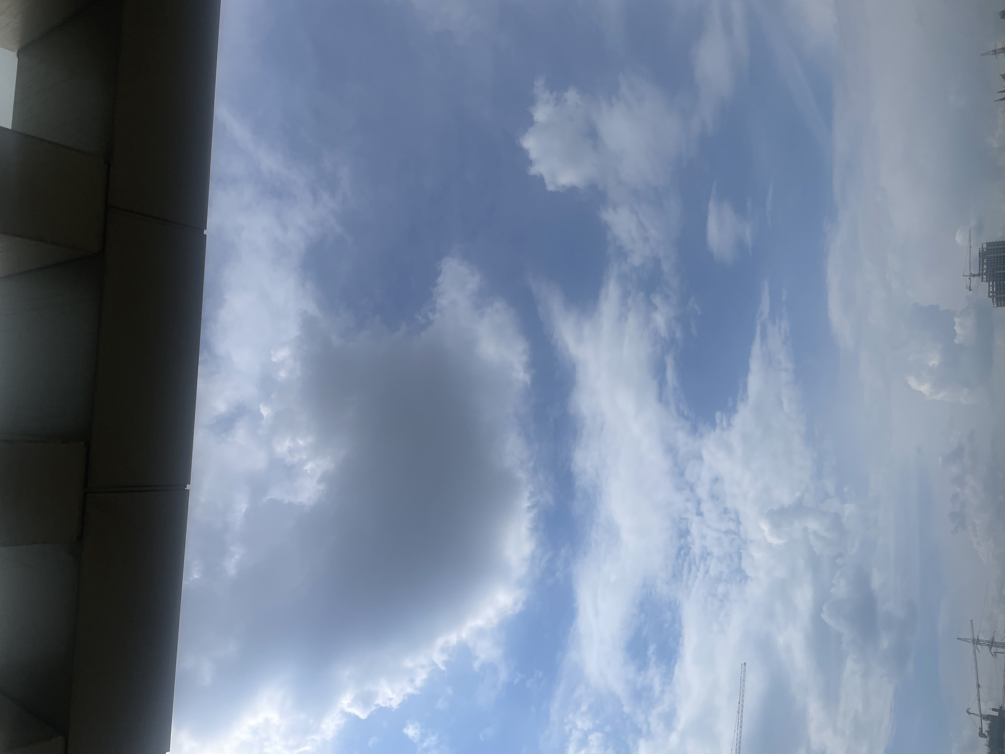 The image, when oriented correctly (landscape), showcases a wide expanse of a partly cloudy sky above an urban environment in Yerevan, Armenia.
On the far left, a significant portion of a dark-grey architectural structure, likely an overhang or part of a building's exterior, is visible. It comprises angular, rectangular sections, casting deep shadows and providing a strong geometric contrast to the organic shapes of the clouds.
The sky is the dominant subject, filled with various cloud formations. A large, dense, and darker grey cloud mass occupies the central-lower-left area, suggesting more substantial cloud cover. Throughout the rest of the frame, wispy, white, and lighter grey clouds are scattered across the blue sky, some appearing as elongated streaks (cirrus or altocumulus), while others are more diffuse. The blue of the sky ranges from a bright, clear blue to a lighter, more diffused shade where the clouds are thinner.
In the far distance on the right, against the cloudy sky, elements of an urban landscape are discernible. Several tall construction cranes and the incomplete structures of buildings indicate an active construction site or a developing city area. These distant features are somewhat indistinct due to atmospheric haze or distance.
The scene depicts daytime conditions with a mix of clear blue sky and considerable cloud cover, suggesting either an approaching change in weather or a typical partly cloudy day. There are no people visible, and no specific event or activity is taking place beyond the ongoing construction. No visible text is present in the image.